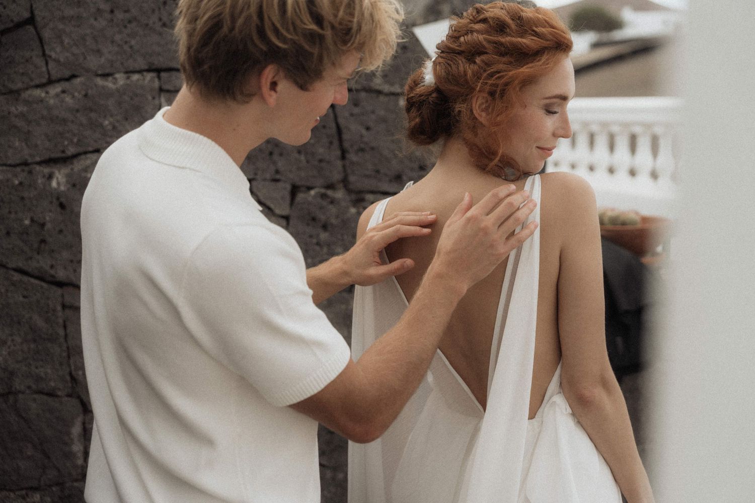 A man in a white shirt fastens the back of a womans elegant white wedding dress outdoors.