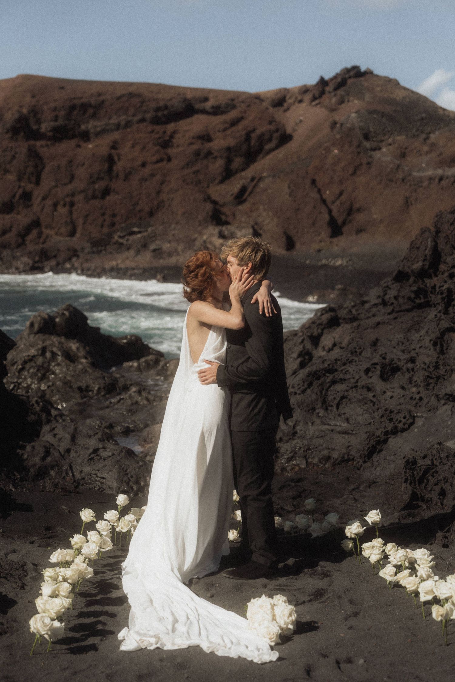 Bride and groom share first kiss on black sand beach with volcanic rocks and white roses.