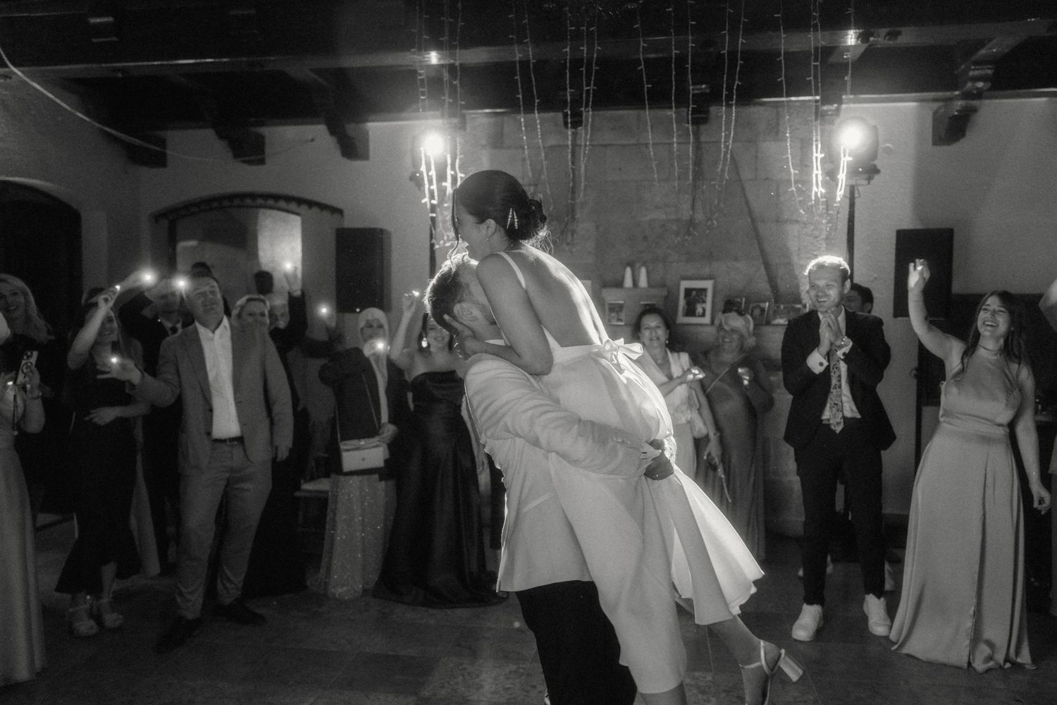 Groom lifting bride on dance floor surrounded by cheering guests in black and white.