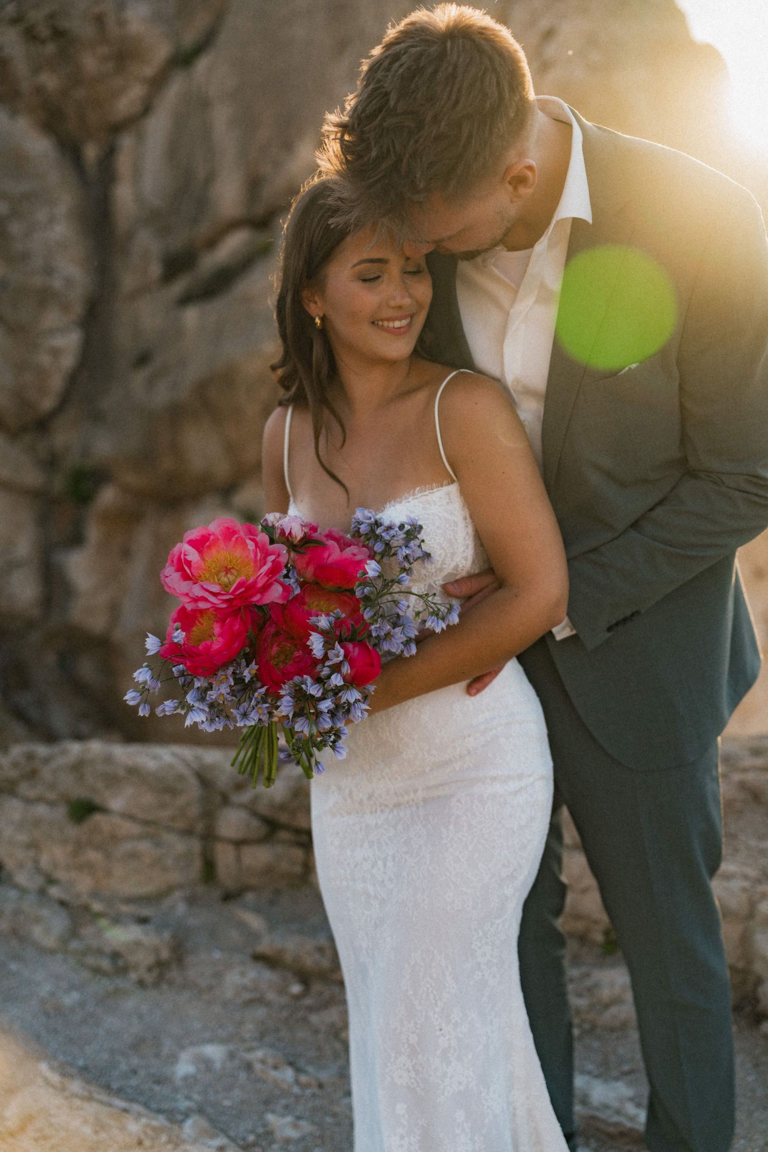 Bride smiles holding vibrant pink peony bouquet as groom kisses her head at sunset near rocky cliffs.