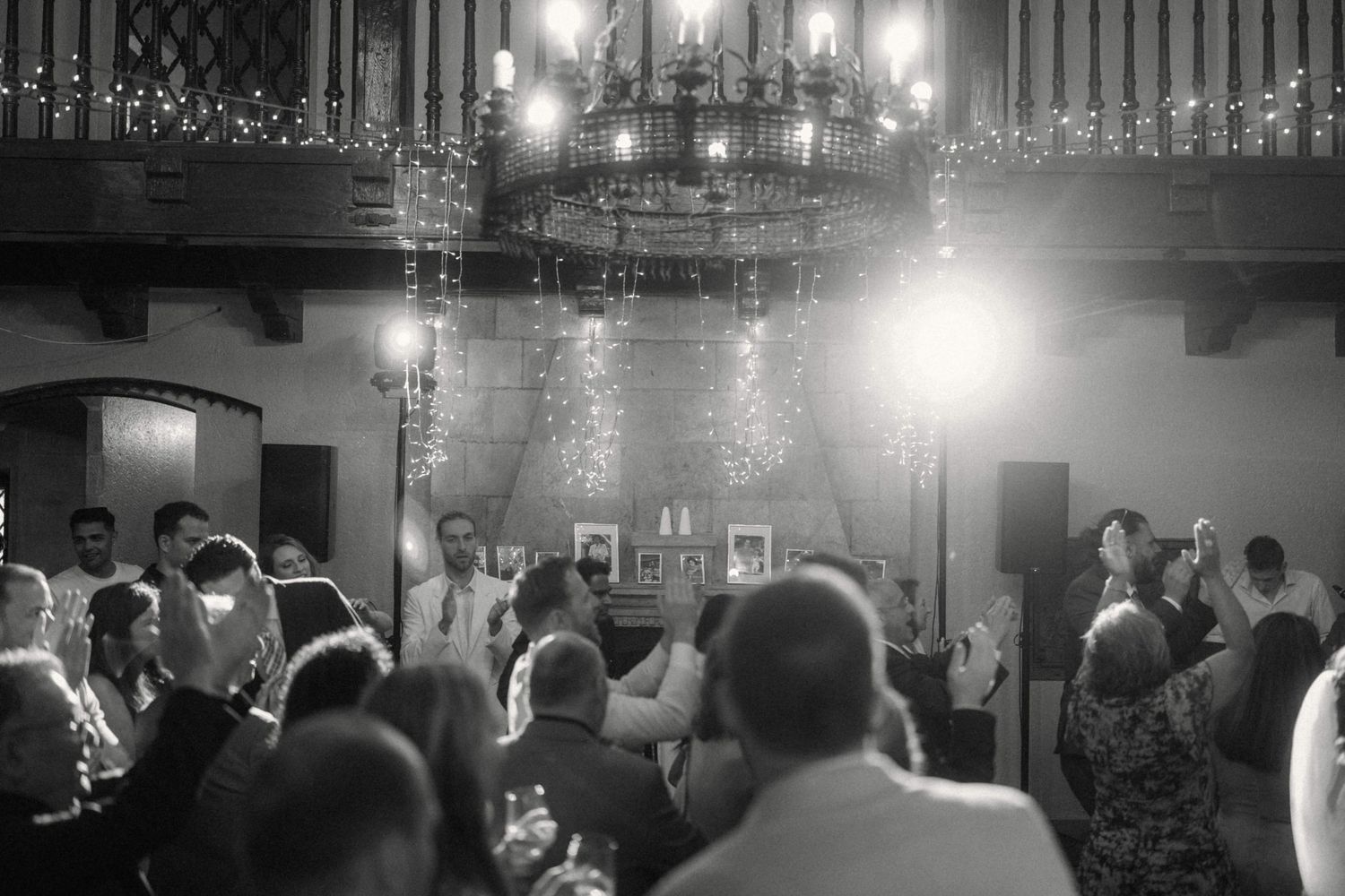 Black and white wide shot of wedding reception guests gathered under chandelier with fairy lights.