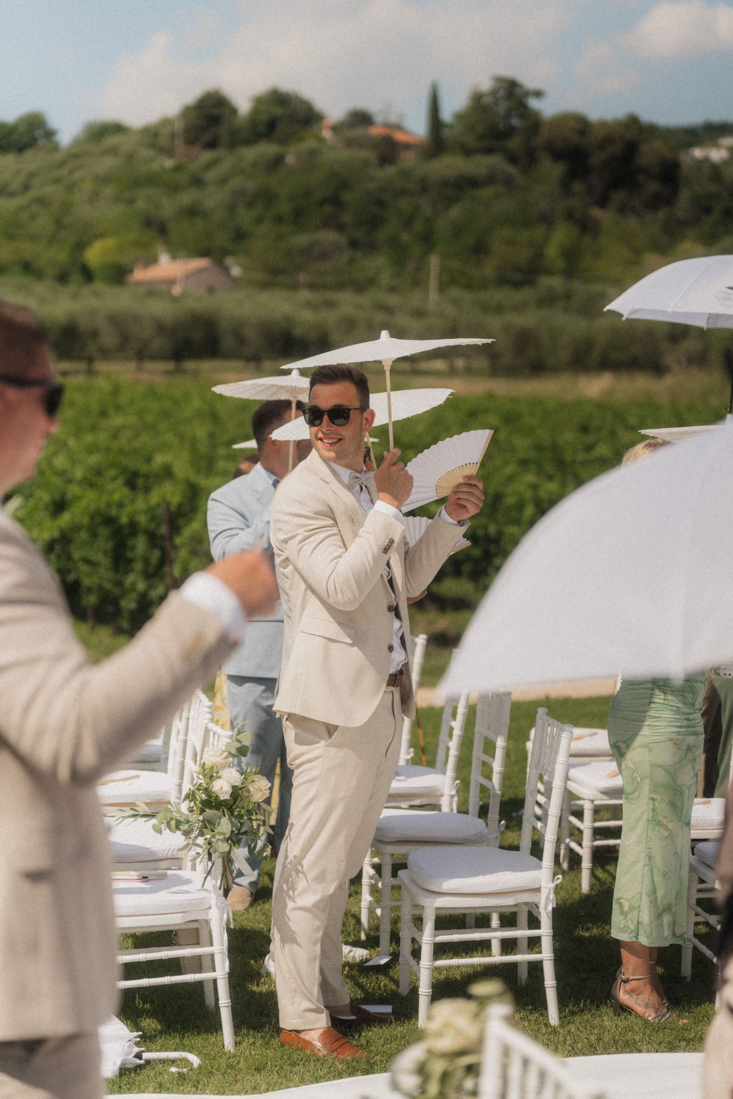 Groomsman in beige suit holding white parasol at outdoor vineyard wedding ceremony on sunny day.