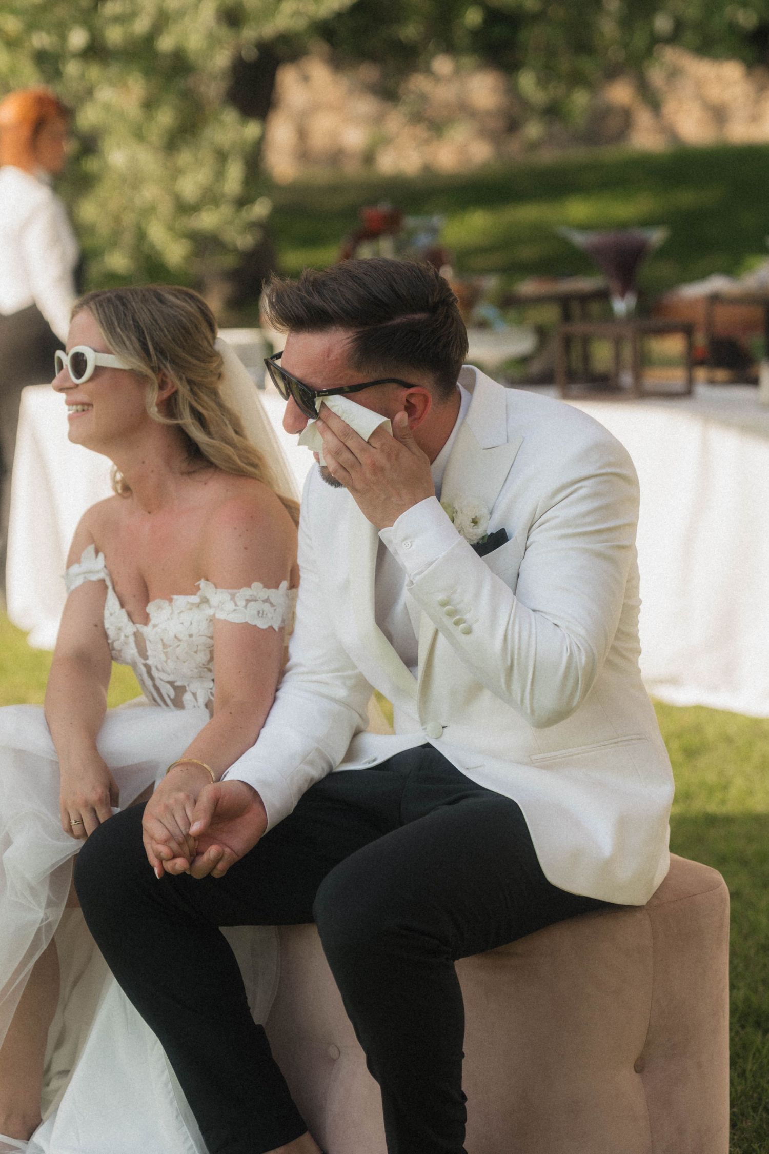 Bride and groom sitting outdoors in sunglasses, groom wiping tears at wedding reception.