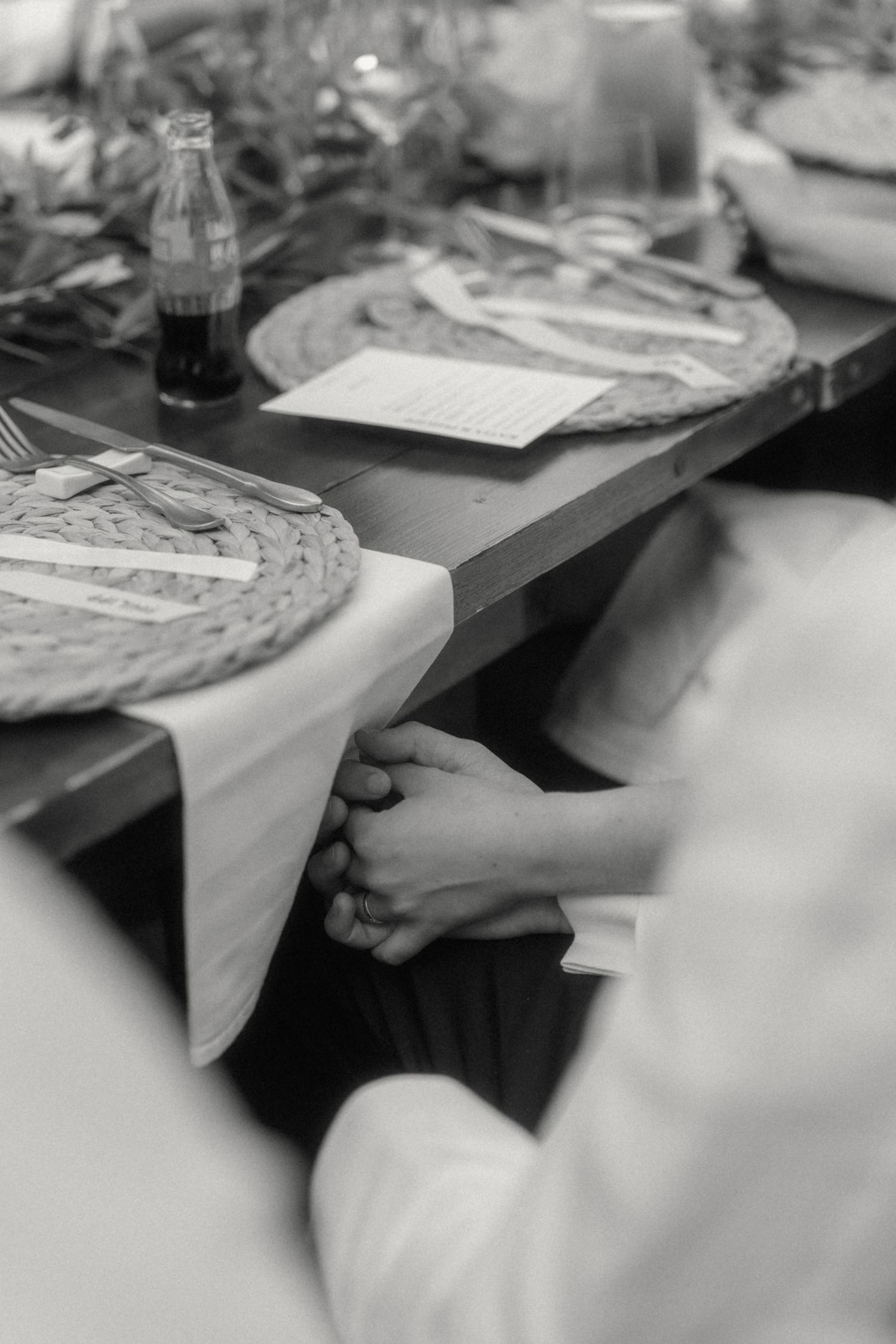 Black and white photo of couple holding hands under decorated wedding reception table with woven placemats.