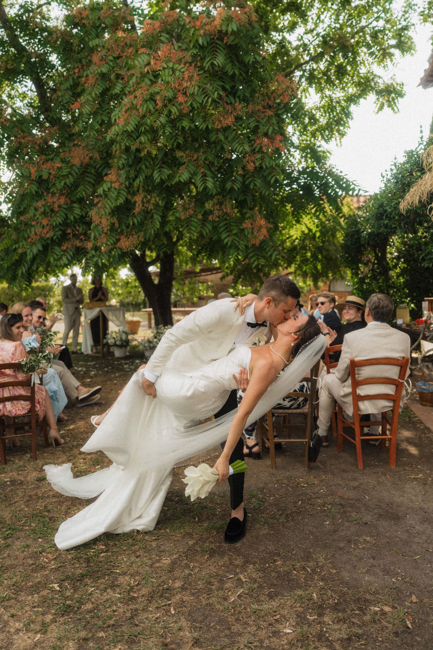 Groom dips bride for a romantic kiss under a flowering tree at outdoor wedding ceremony.