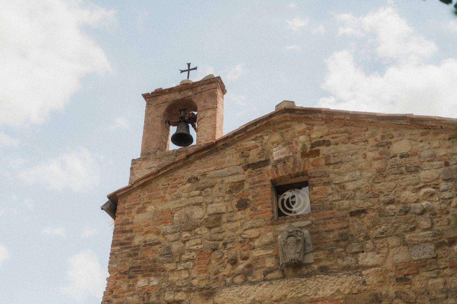 Ancient stone church bell tower with cross against cloudy sky in Tuscany, Italy.