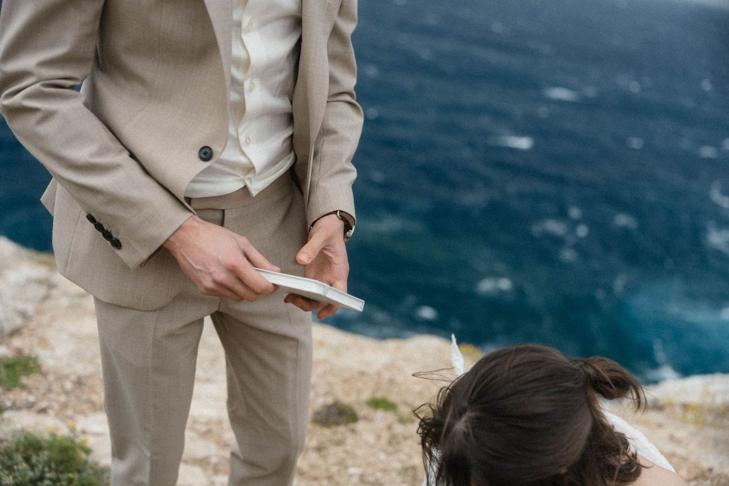 Groom in beige suit holding wedding vows booklet on rocky cliffside with blue ocean backdrop.
