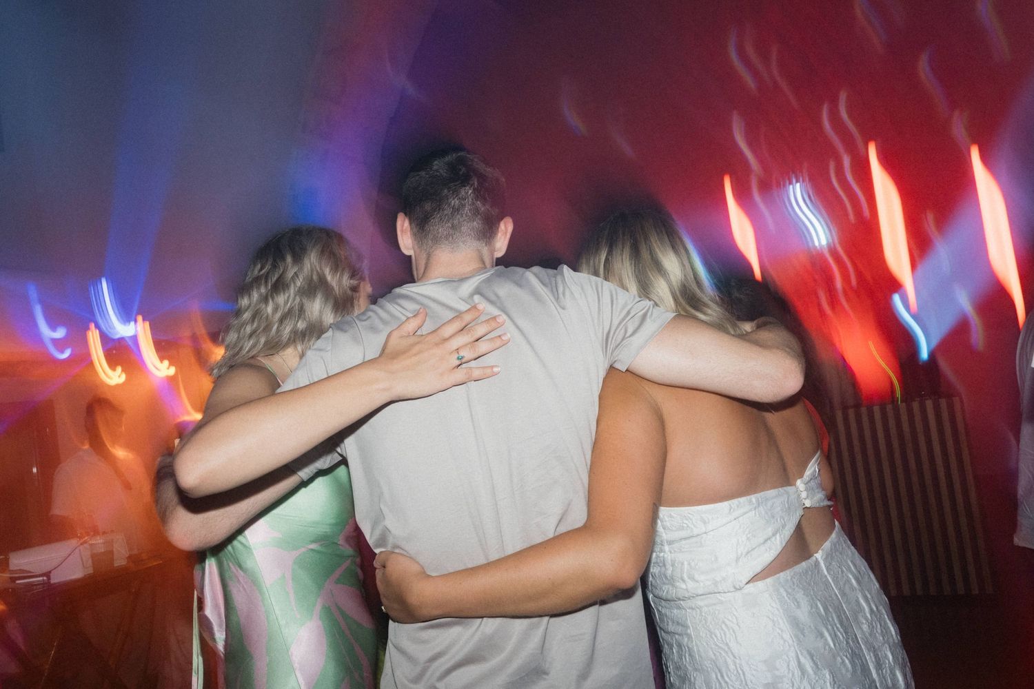 Three friends embrace on a dance floor surrounded by colorful red and blue party lights at a nightclub event.