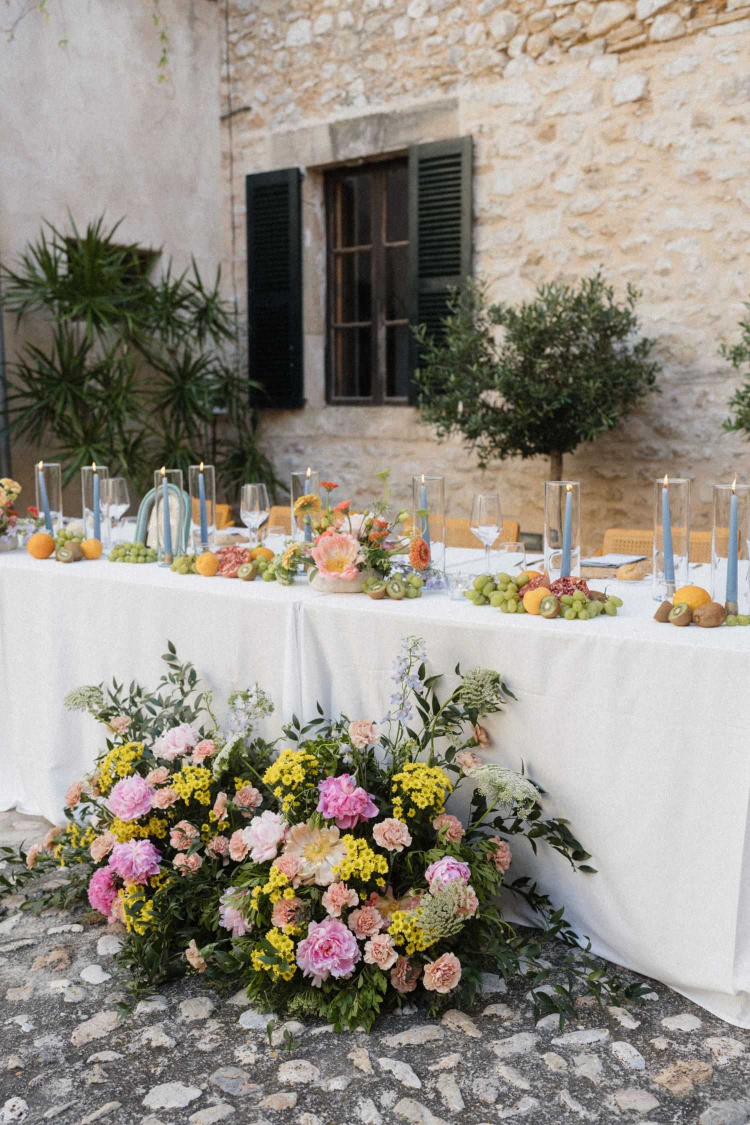 Outdoor wedding reception table adorned with colorful flowers, fruit, and candles against stone wall.
