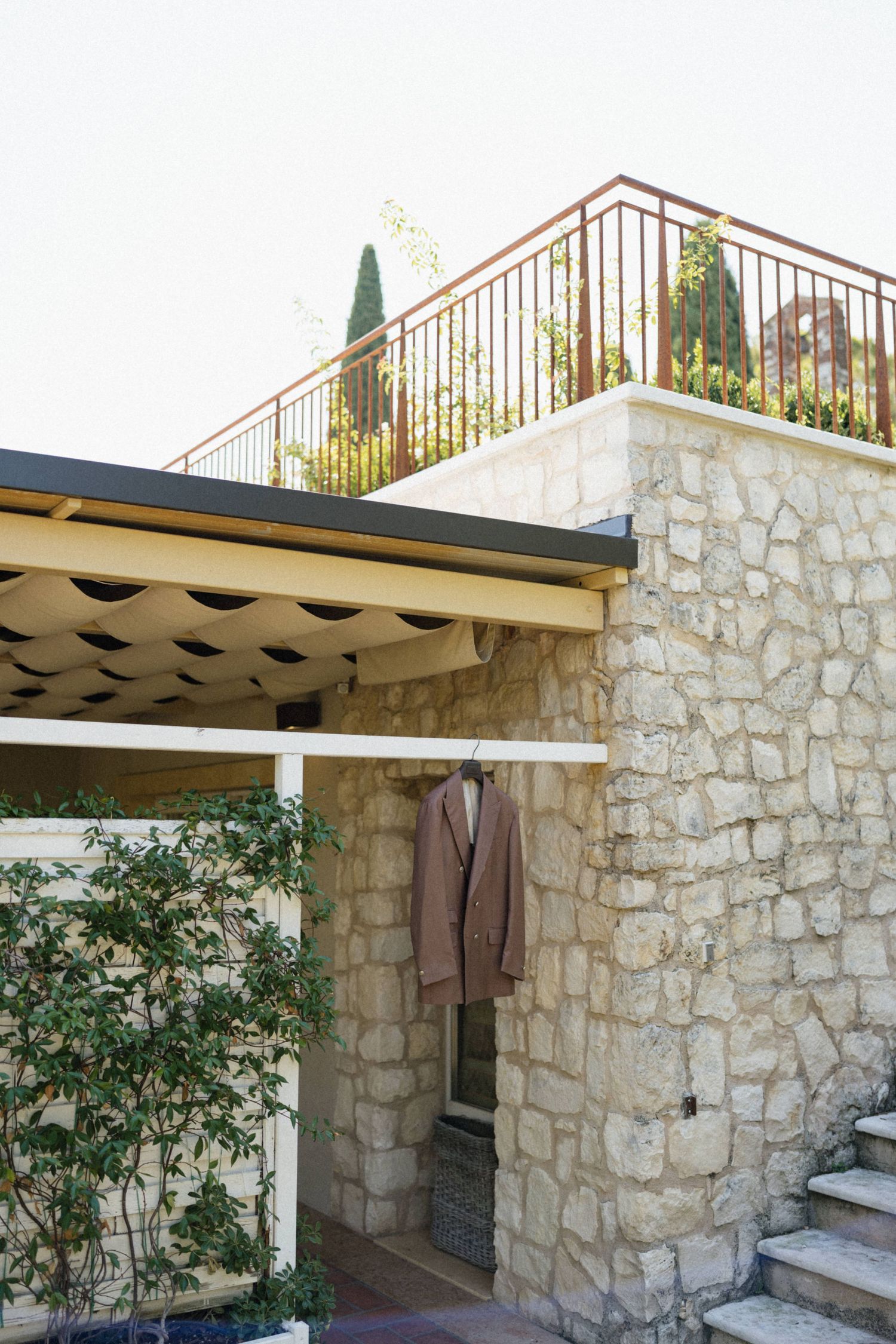 Stone building exterior with tan blazer hanging on wall, upper balcony with iron railing and cypress trees.