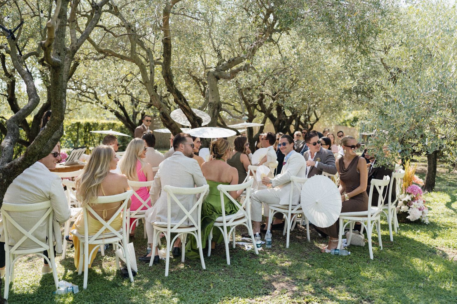 Guests gathered around white chairs at outdoor tables under olive trees for an elegant garden wedding reception.