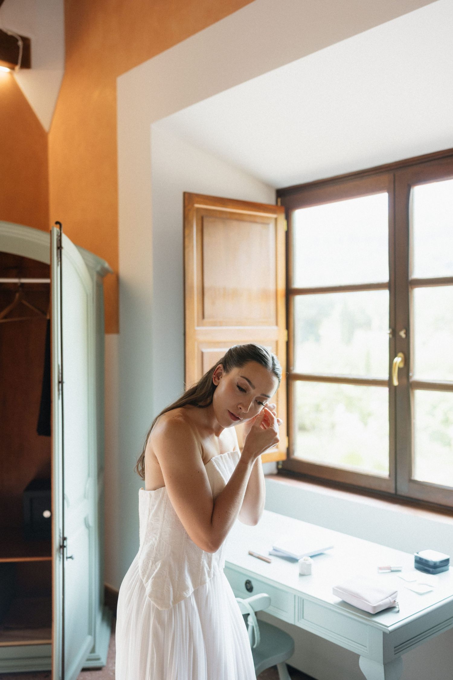 Bride in a white strapless wedding dress puts on earrings beside a sunlit window in a rustic Tuscan-style bedroom.