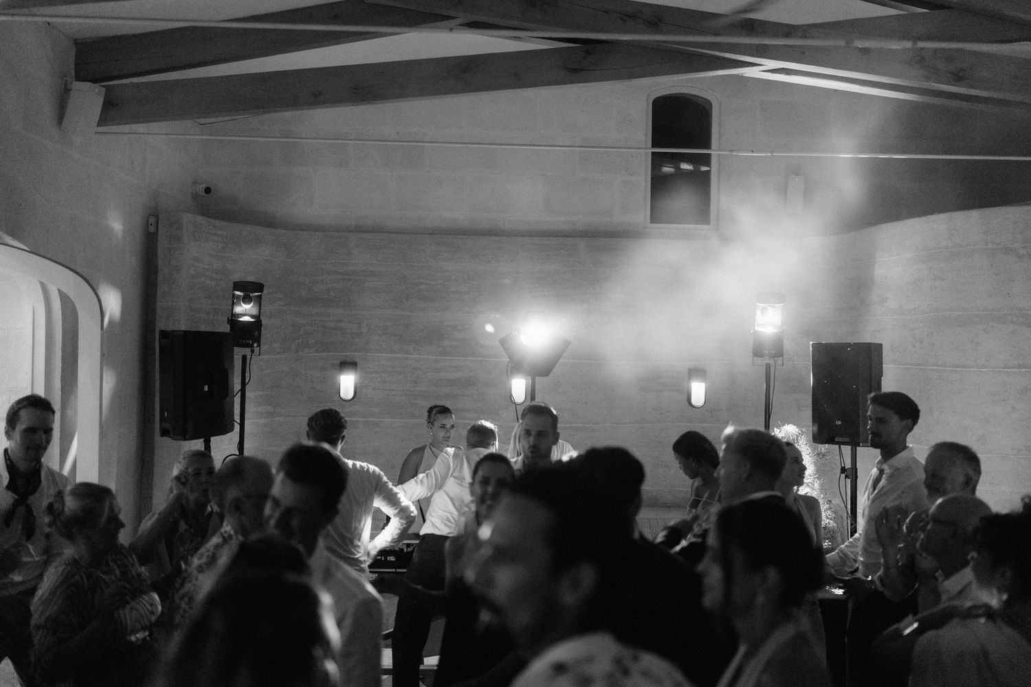 Black and white photo of wedding guests gathered at an evening reception with hazy stage lights and speakers glowing behind them.