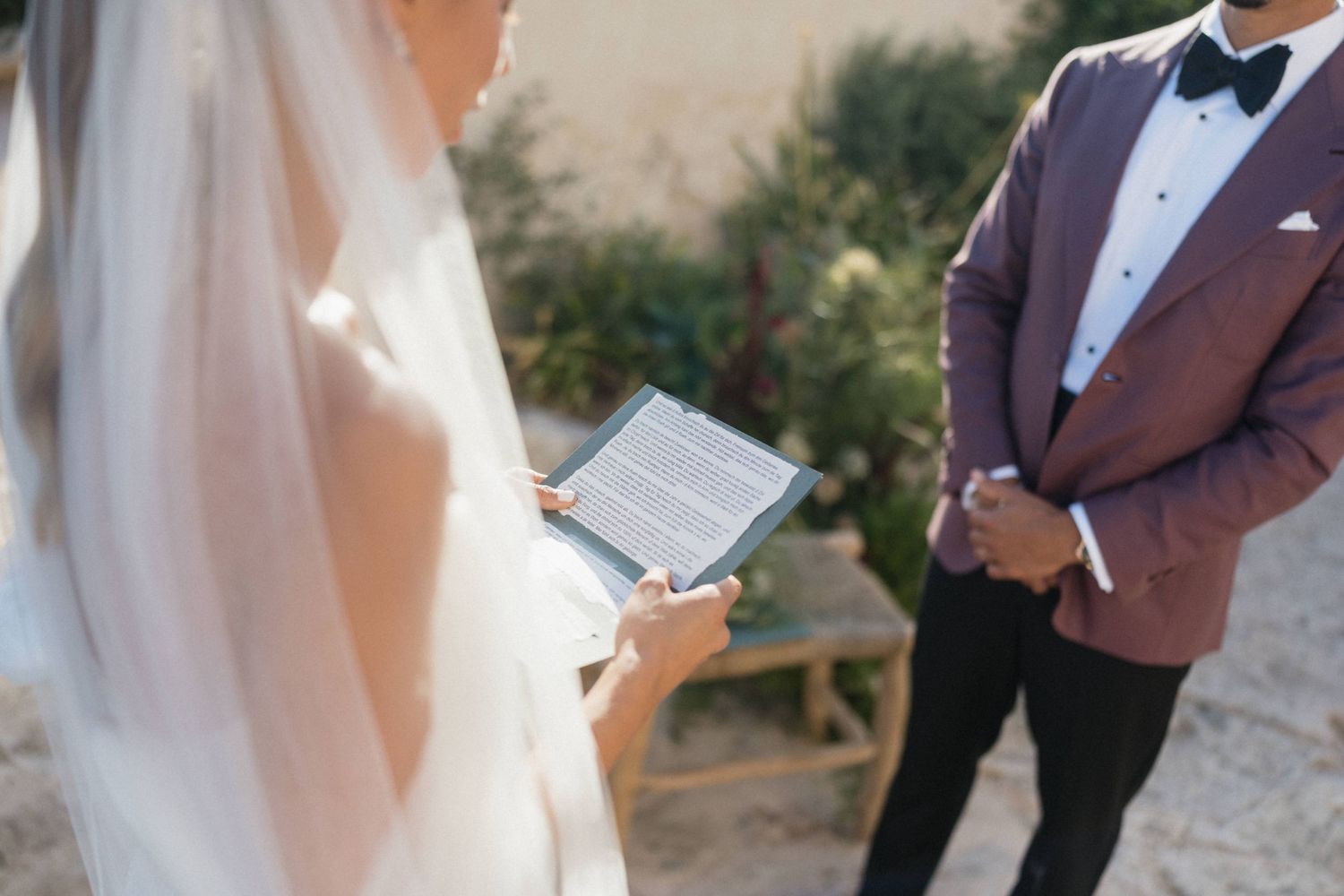Bride in a veil holds a vow book beside a groom in a burgundy blazer and black bow tie during an outdoor ceremony.