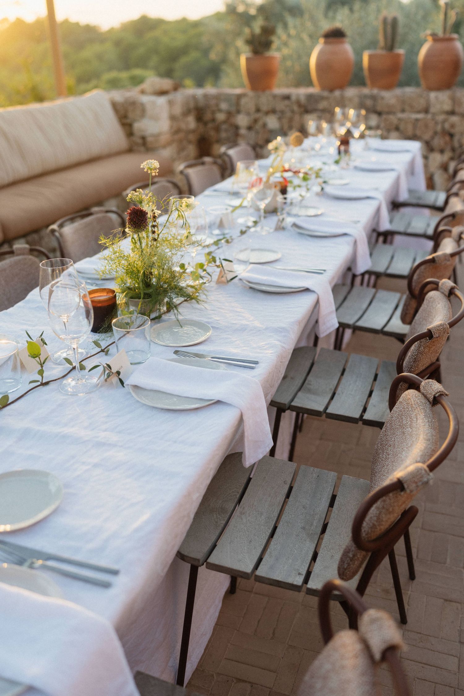 Long wedding reception table set with white linens, wildflower centerpieces, and rustic wooden chairs outdoors.