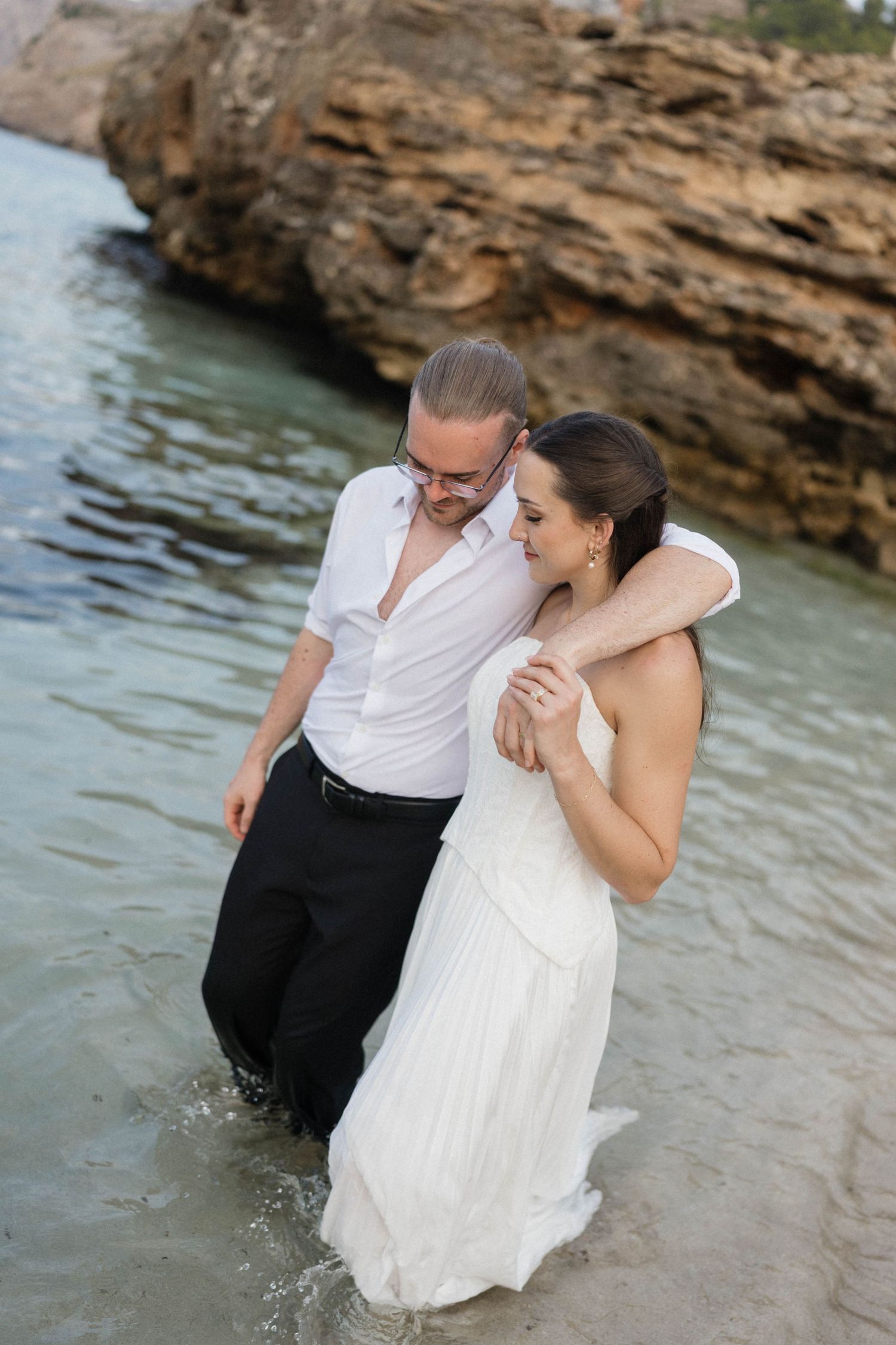Barefoot couple in white wedding attire embracing while wading in shallow turquoise water beside a rocky coastal cliff.