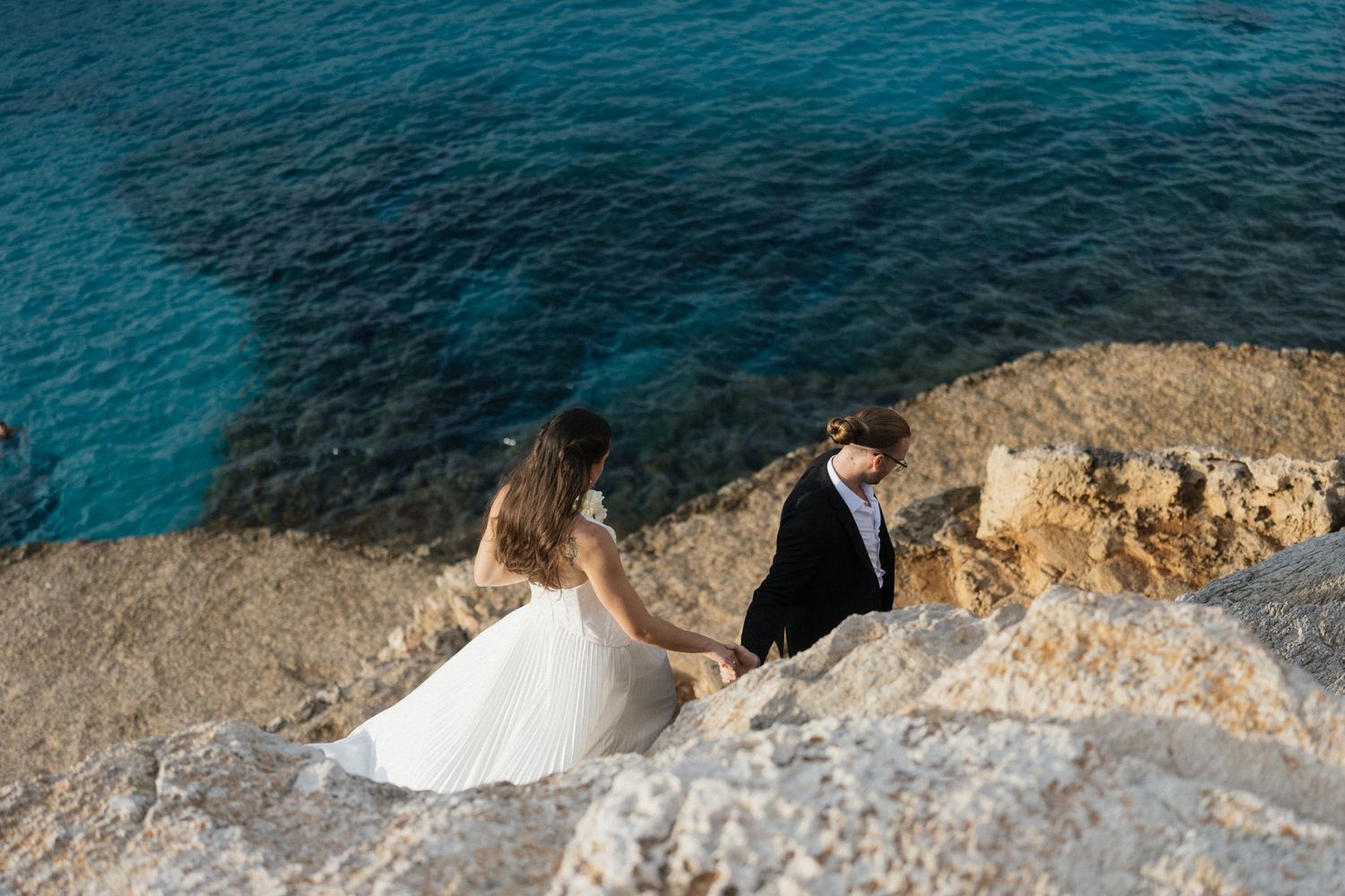 Bride in a white gown and groom in a dark suit walk hand in hand along rocky seaside cliffs above clear turquoise water.