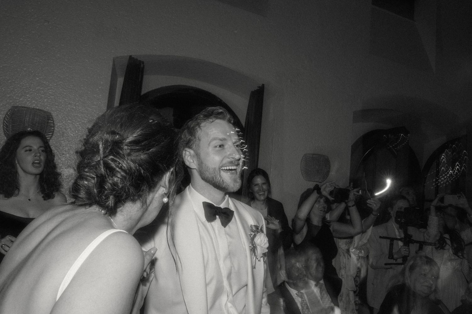 Black and white photo of groom in white tuxedo smiling on dance floor at wedding reception.