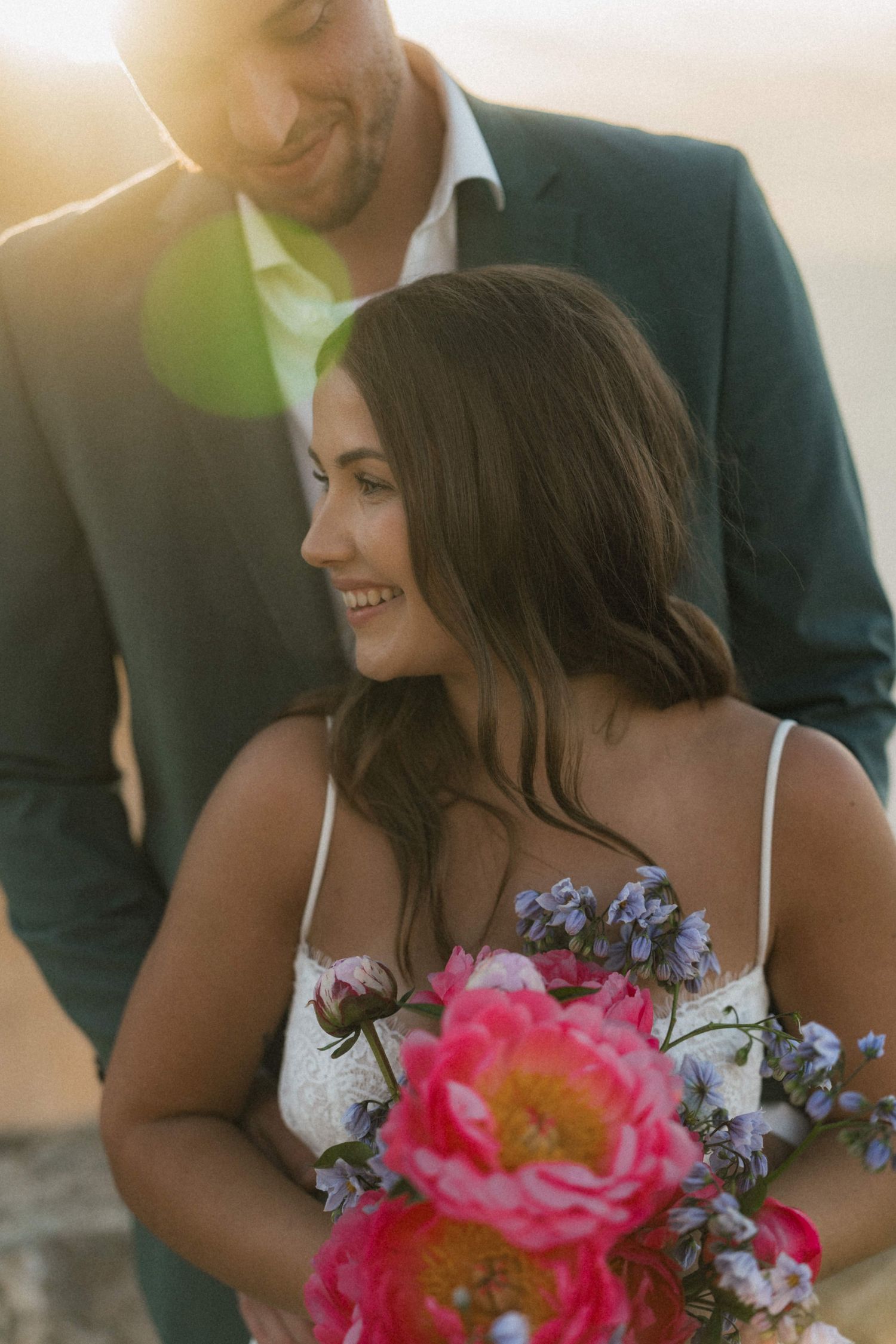 Smiling bride holding vibrant pink peony bouquet with blue flowers, groom in navy suit behind her, golden sunset light.