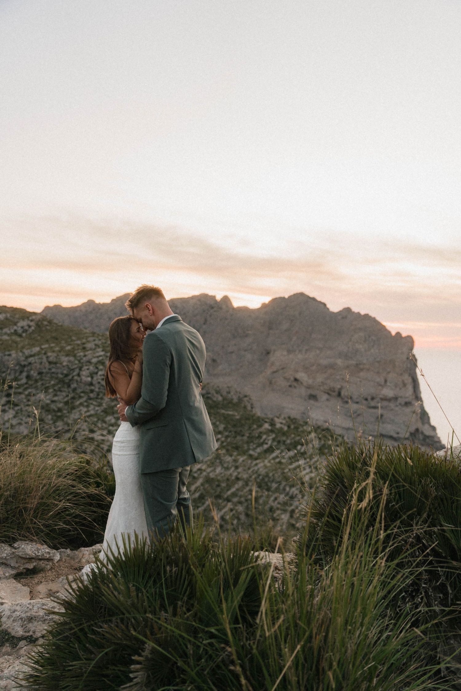 Couple embracing at sunset on a rocky hilltop, groom in grey suit, bride in white fitted gown.