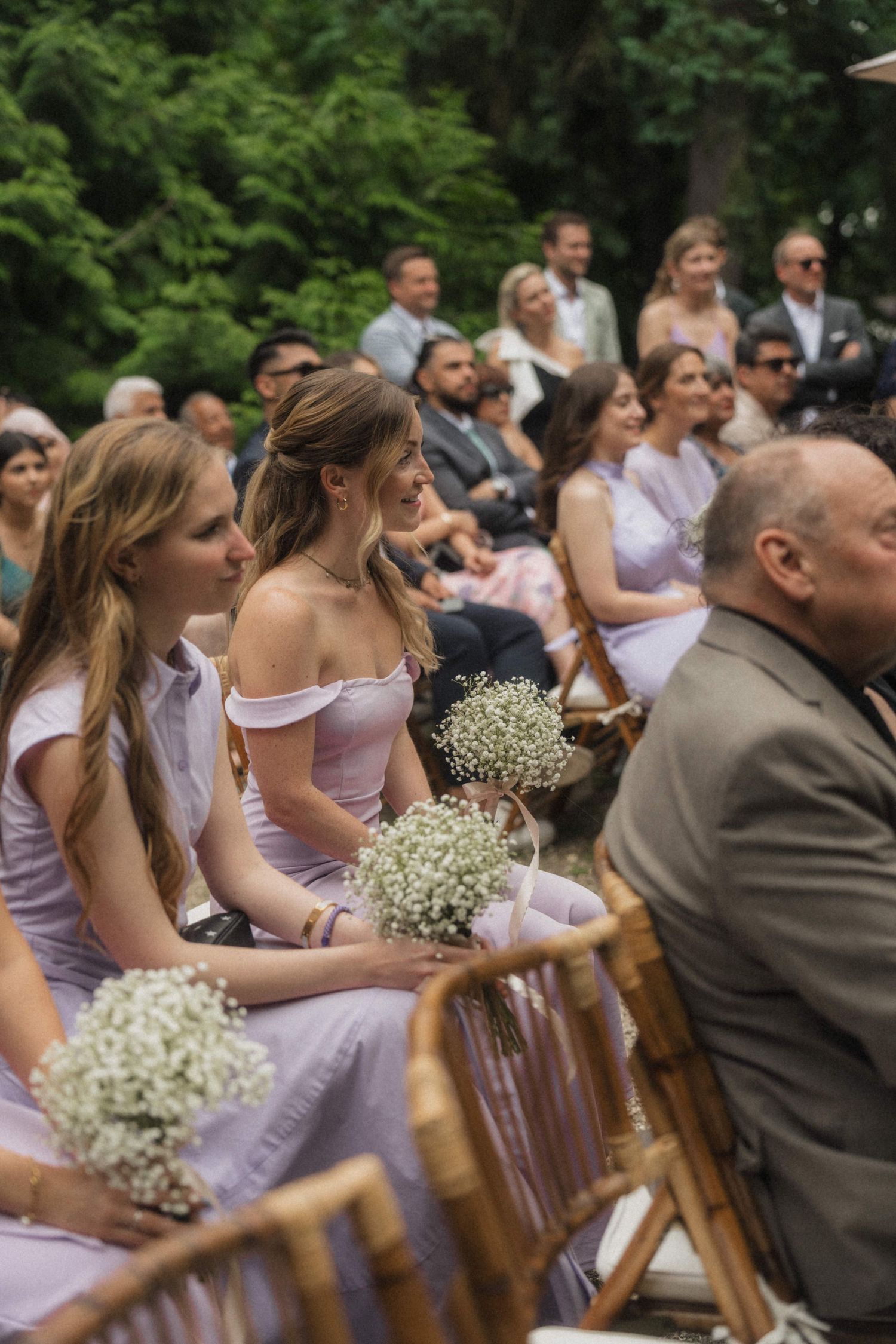 Bridesmaids in lavender dresses holding baby's breath bouquets seated at an outdoor wedding ceremony.