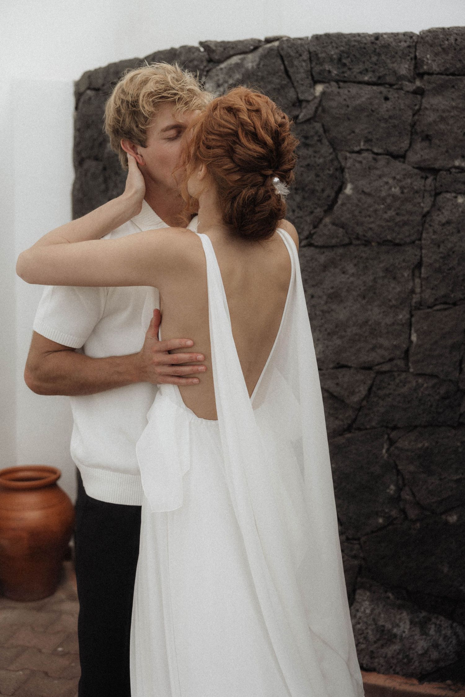 Bride and groom sharing a romantic kiss, bride wearing elegant backless white wedding dress against dark stone wall.