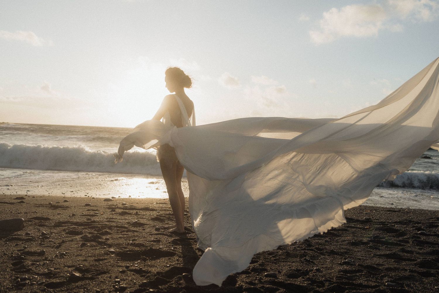 Bride in white gown with billowing veil stands backlit by golden sunset light on a dark sandy beach.