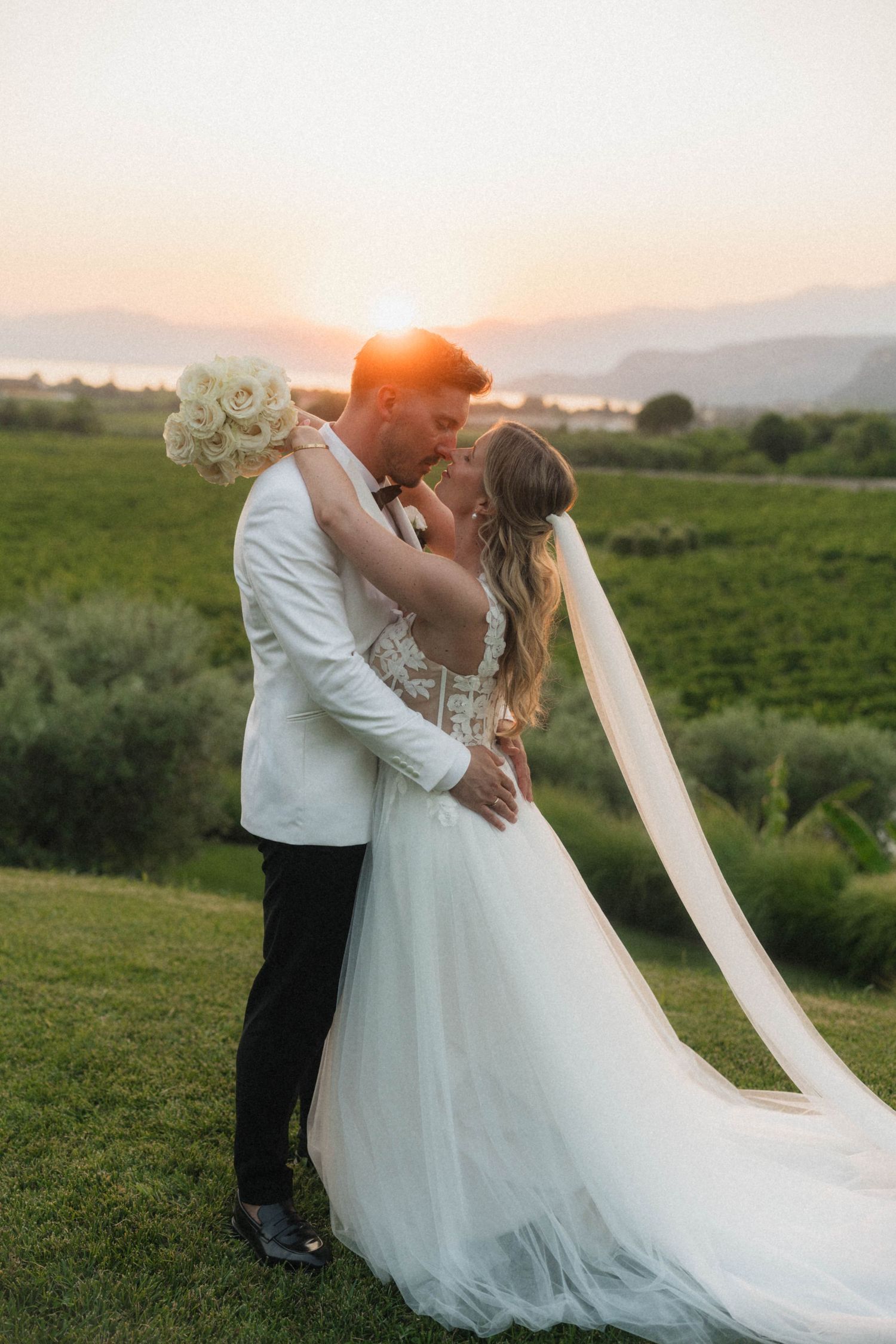 Bride and groom sharing a romantic sunset kiss in a vineyard with mountains and golden light behind them.