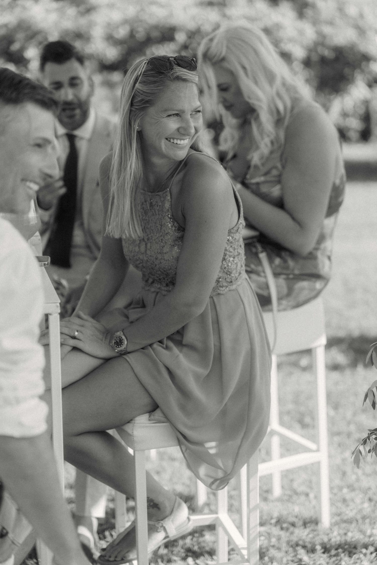 Black and white photo of a smiling blonde wedding guest in a short dress seated outdoors at a garden ceremony.