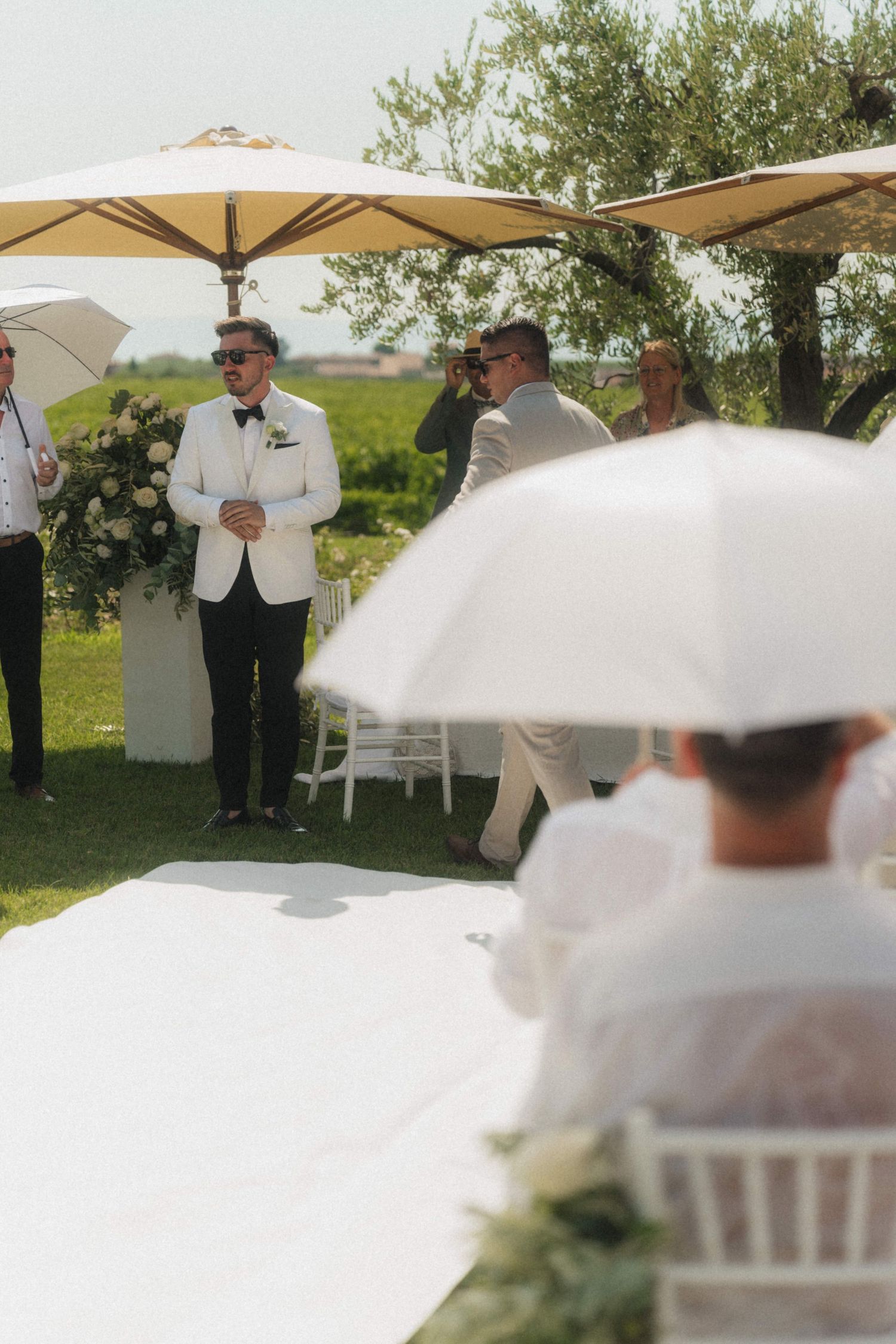 Groom in white tuxedo jacket stands at outdoor wedding ceremony aisle, guests seated under large umbrellas.