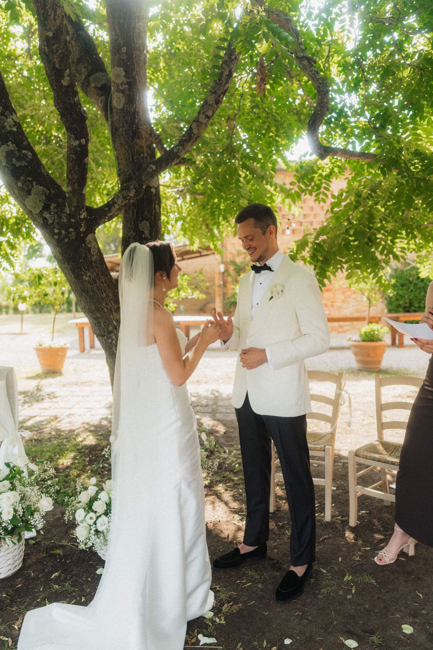 Bride placing wedding ring on groom's finger during outdoor ceremony under lush green tree, both in elegant white attire.
