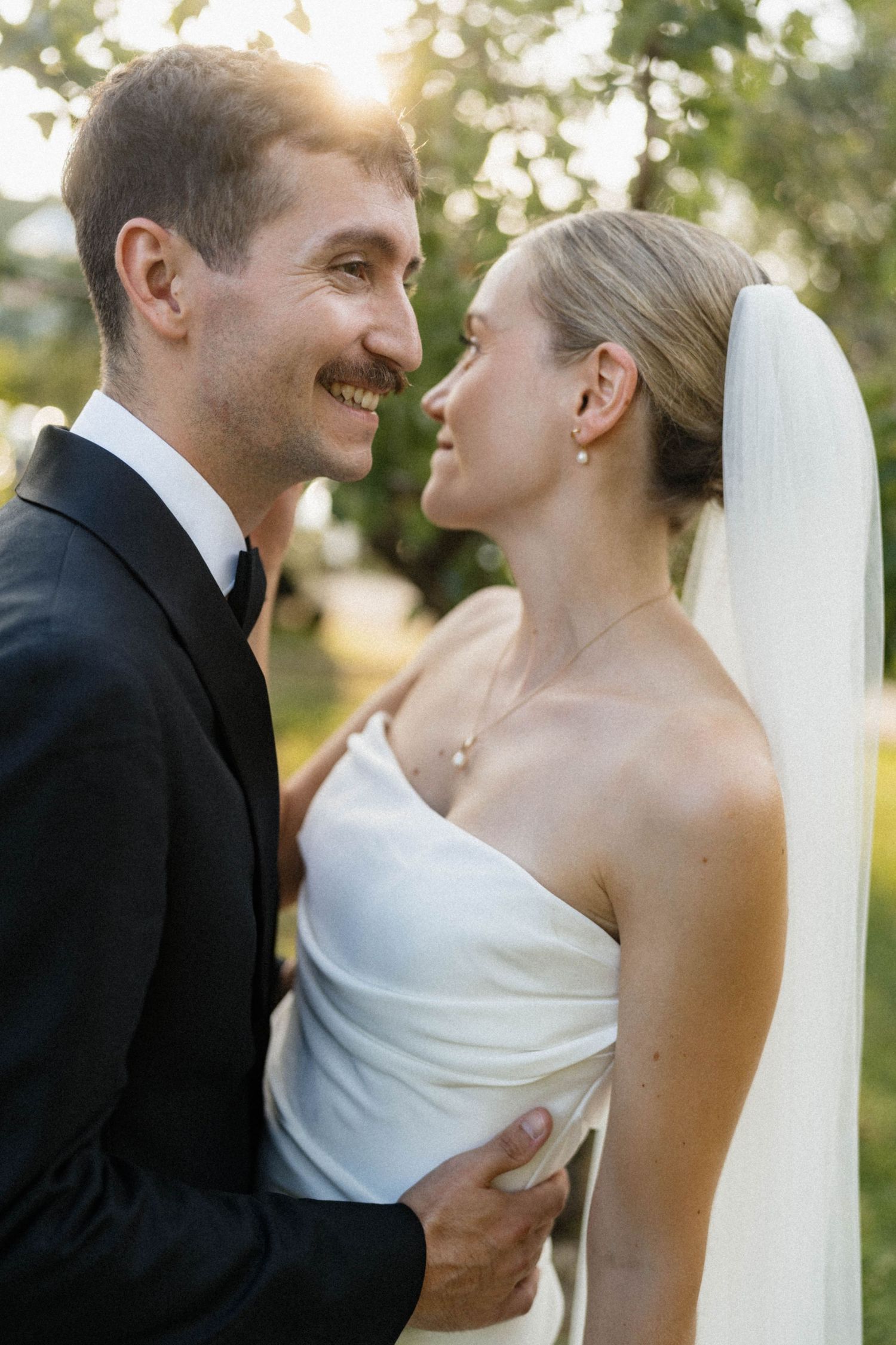 Smiling groom in black tuxedo gazes at bride in strapless white gown during golden hour outdoor wedding portrait.