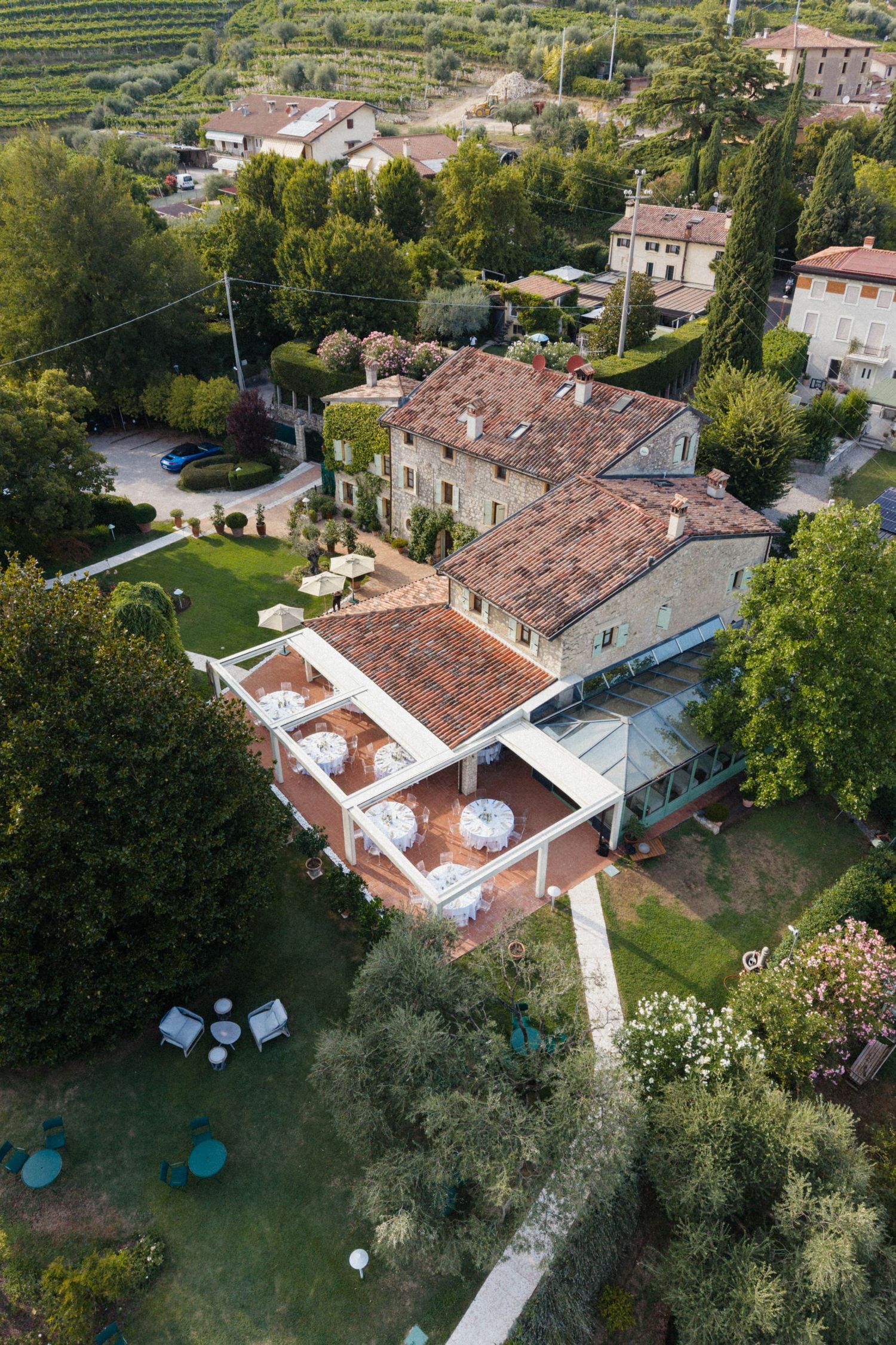 Aerial view of a luxurious Italian villa with terracotta roof, manicured gardens, and outdoor dining terrace surrounded by lush trees.