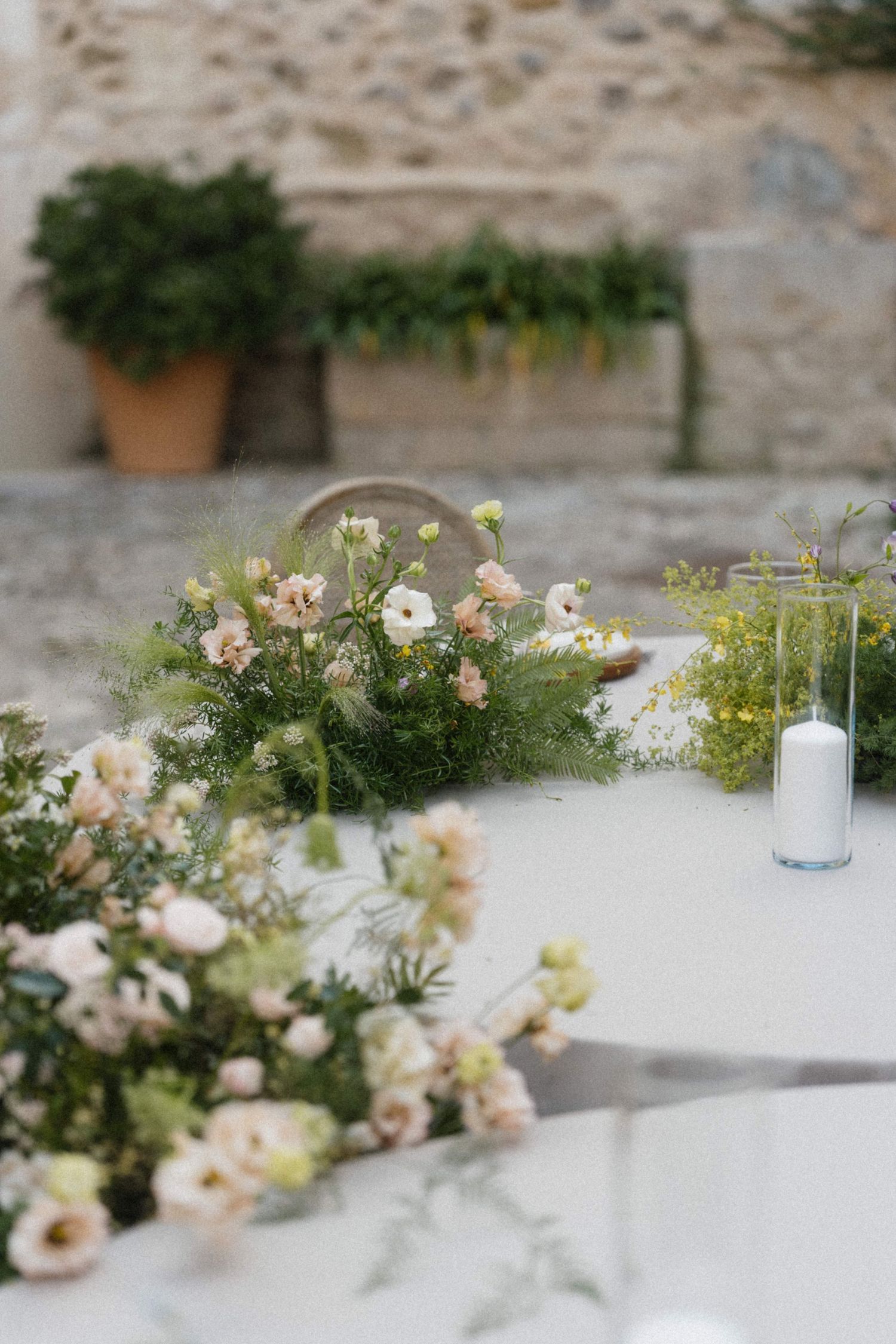Elegant outdoor wedding table with blush pink flowers, greenery arrangements, and white pillar candles on white linen.