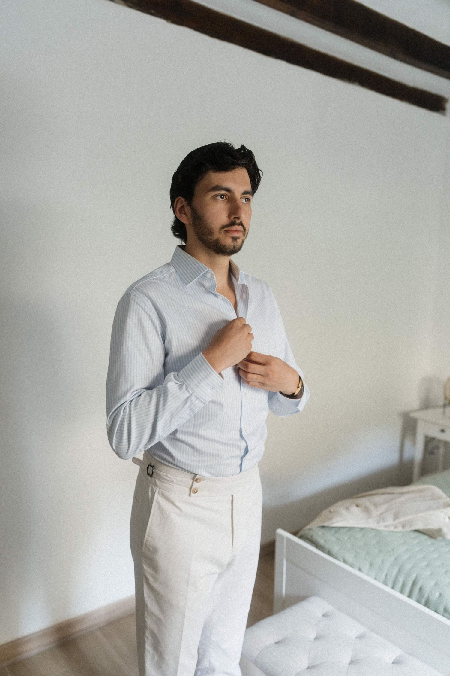 Groom buttoning his light blue dress shirt while getting ready in a bright bedroom before his wedding.