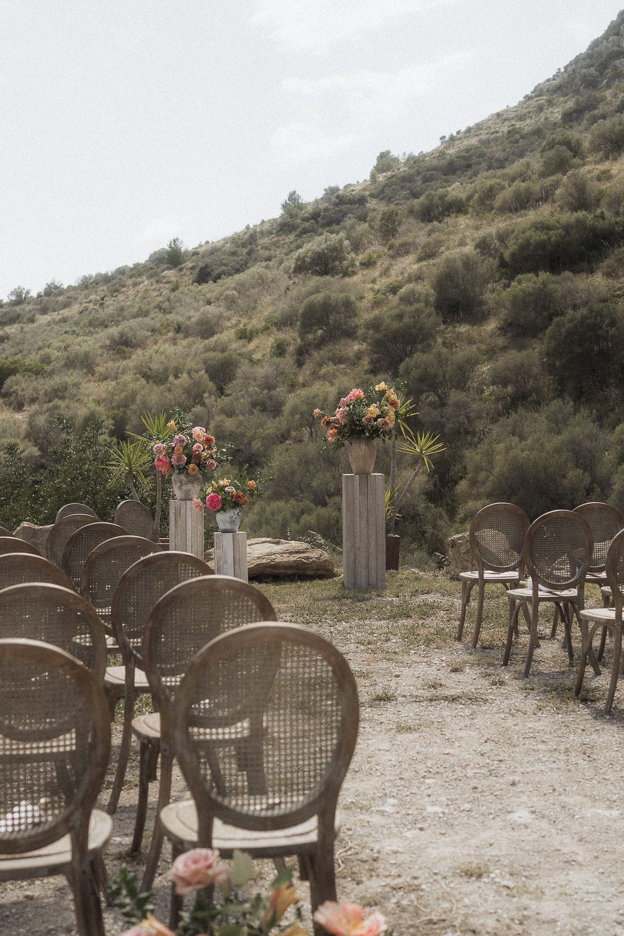 Outdoor wedding ceremony setup with rattan chairs lining a gravel aisle, floral arrangements on pedestals, and a hillside backdrop.