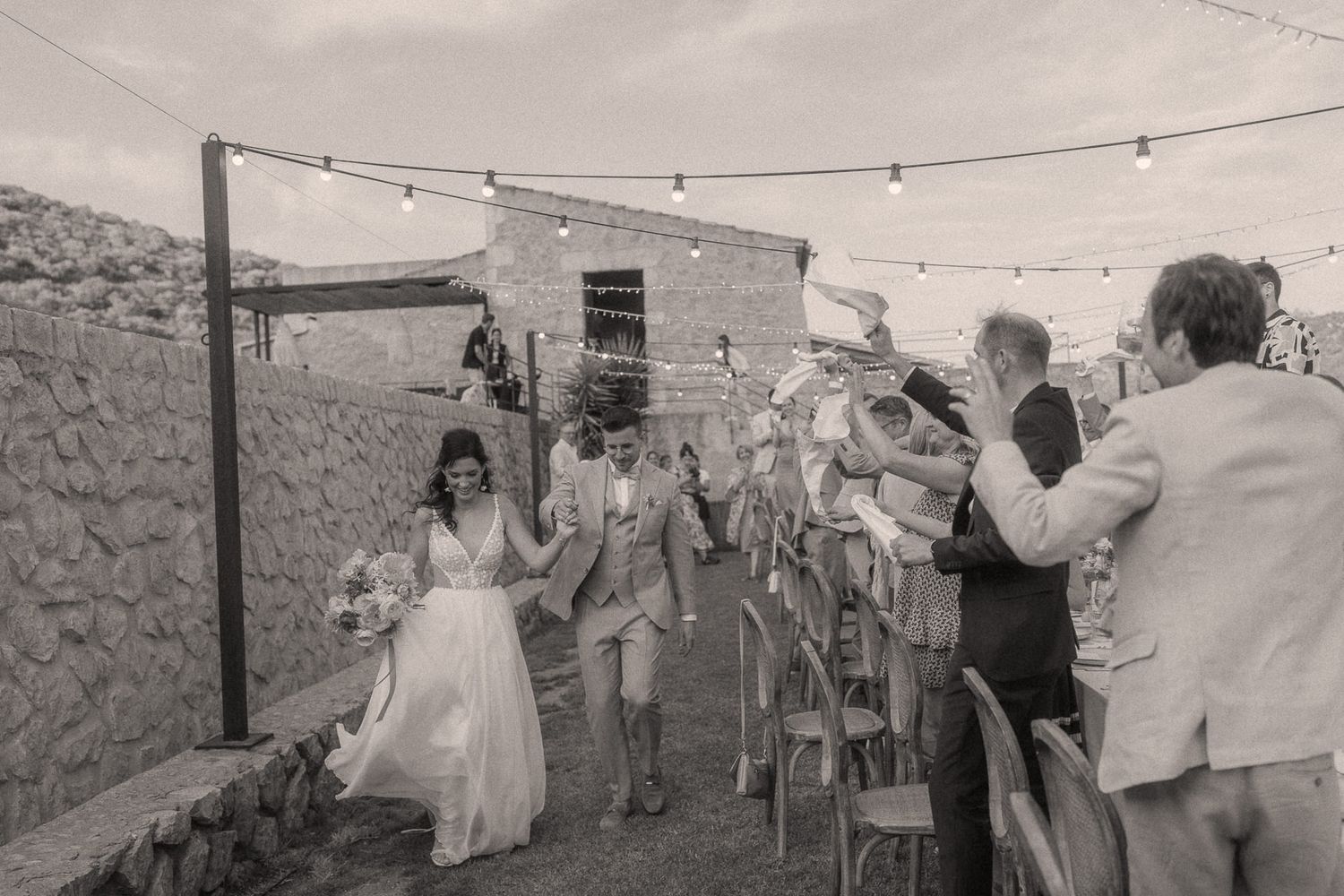 Black and white photo of newlyweds walking through cheering guests at outdoor wedding venue with string lights.