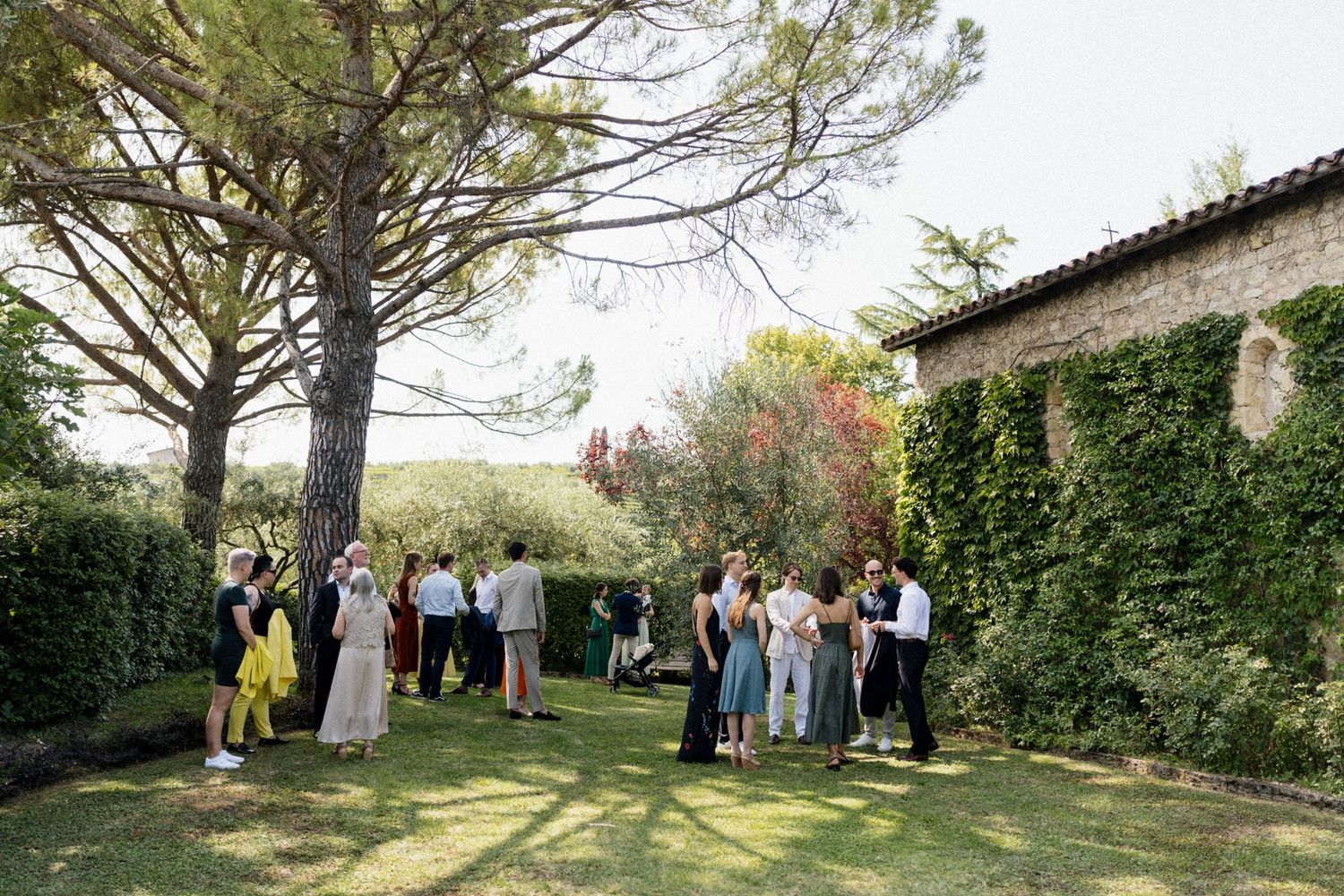 Guests mingle on a lush green lawn beside a stone villa surrounded by tall pine trees at an outdoor garden party.