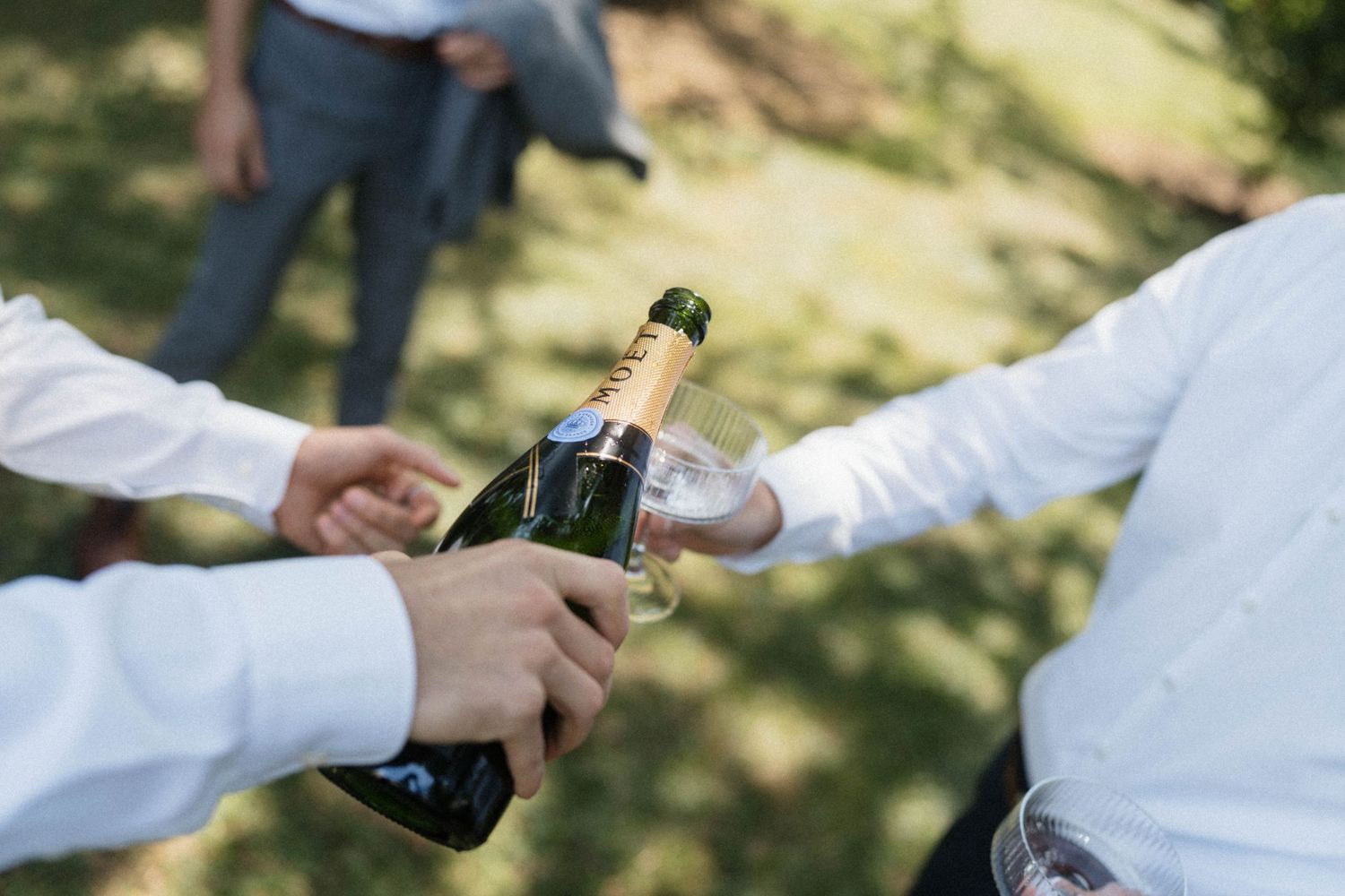 Close-up of hands opening a champagne bottle outdoors at a wedding celebration in sunlight.
