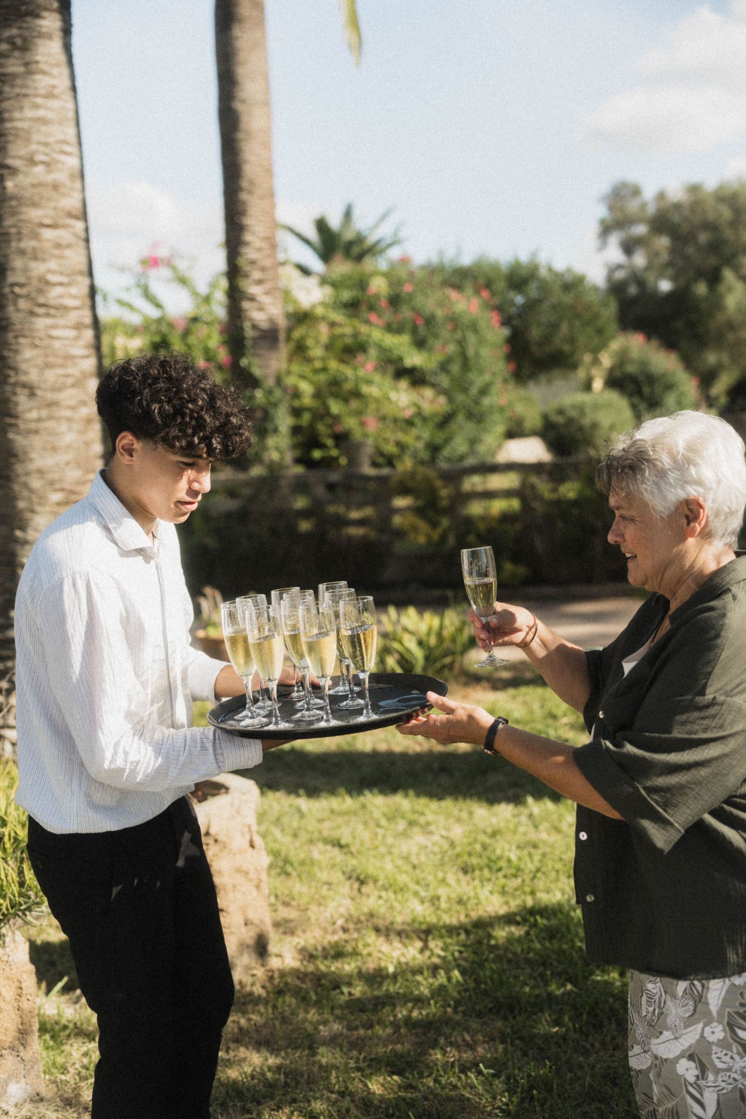 Young waiter serving champagne flutes on a tray to elderly wedding guest outdoors.
