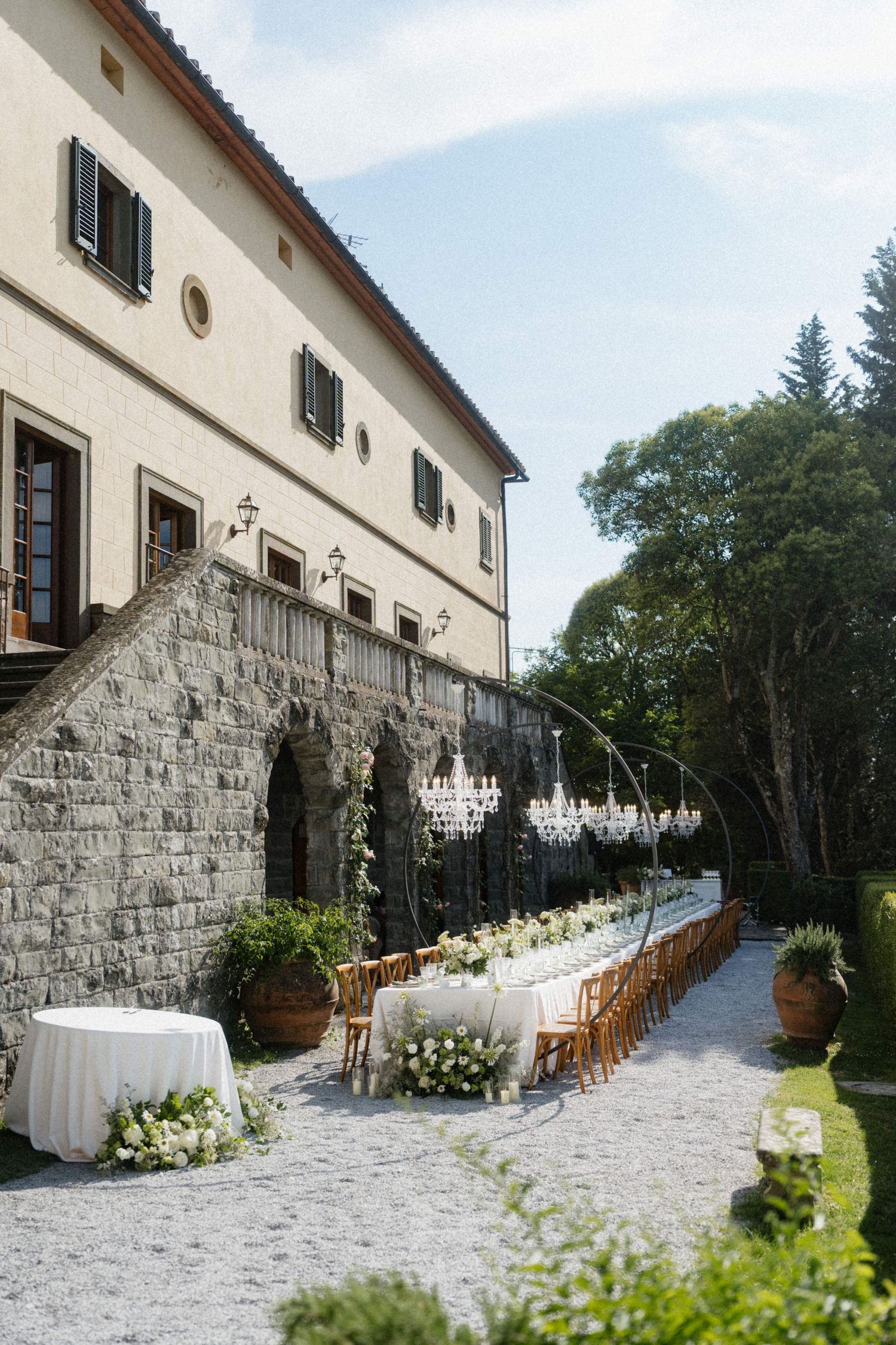 Outdoor Italian villa wedding reception with long dining table, crystal chandeliers, and lush garden surroundings.