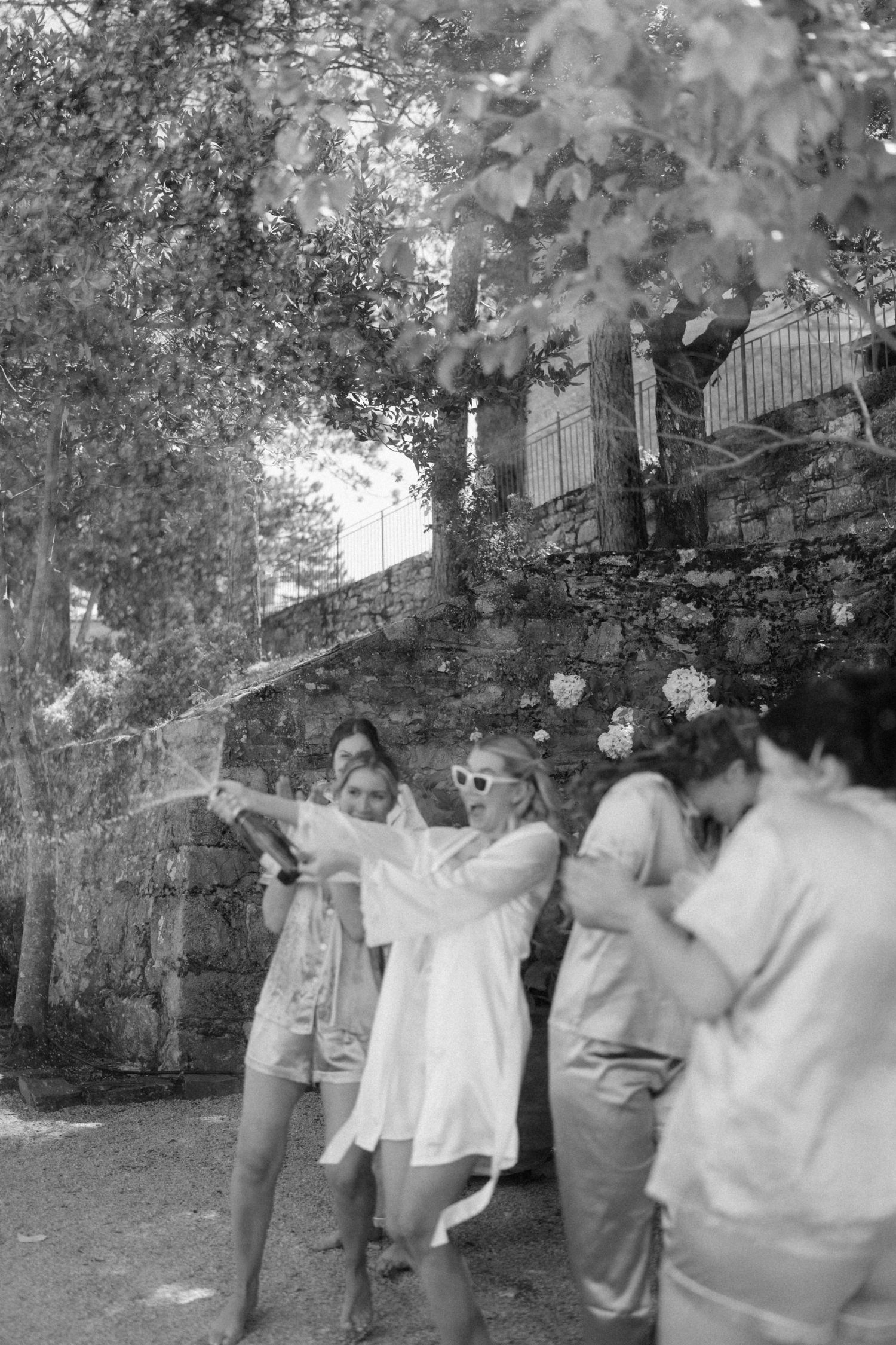 Black and white photo of bridesmaids popping champagne outdoors near stone wall and trees.