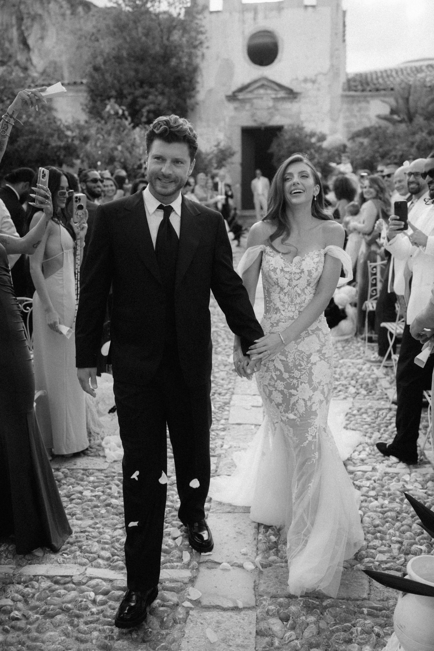 Black and white photo of smiling newlyweds walking down cobblestone aisle after ceremony.