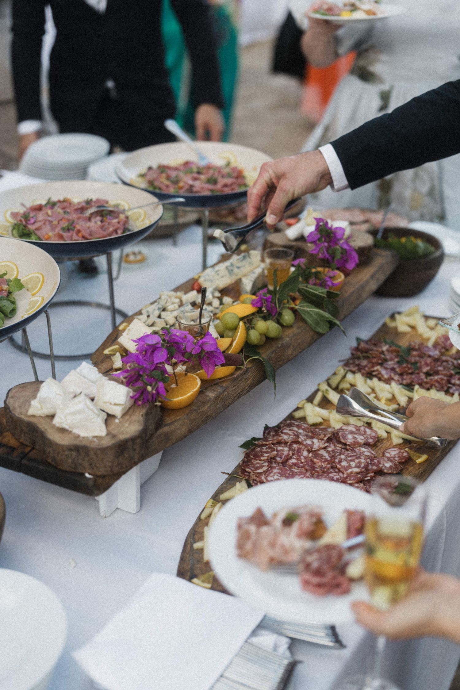 Wedding catering spread with charcuterie, cheese, fruits, and purple flowers on rustic wooden boards at reception.