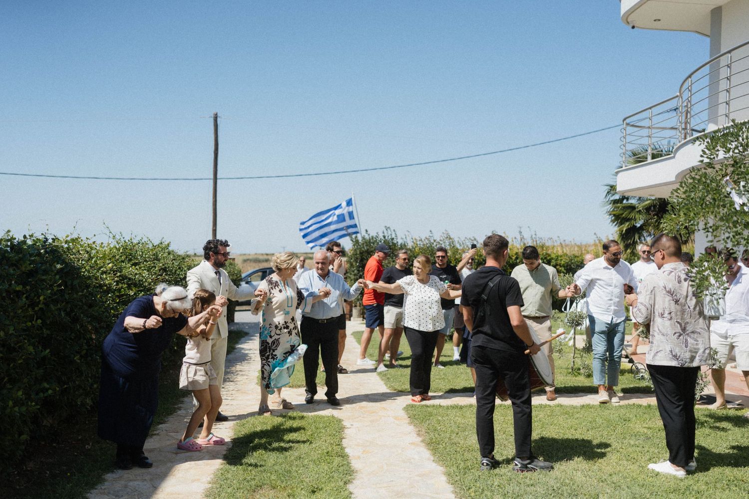 Group of guests gathered outside a white building with a Greek flag during a sunny outdoor celebration.