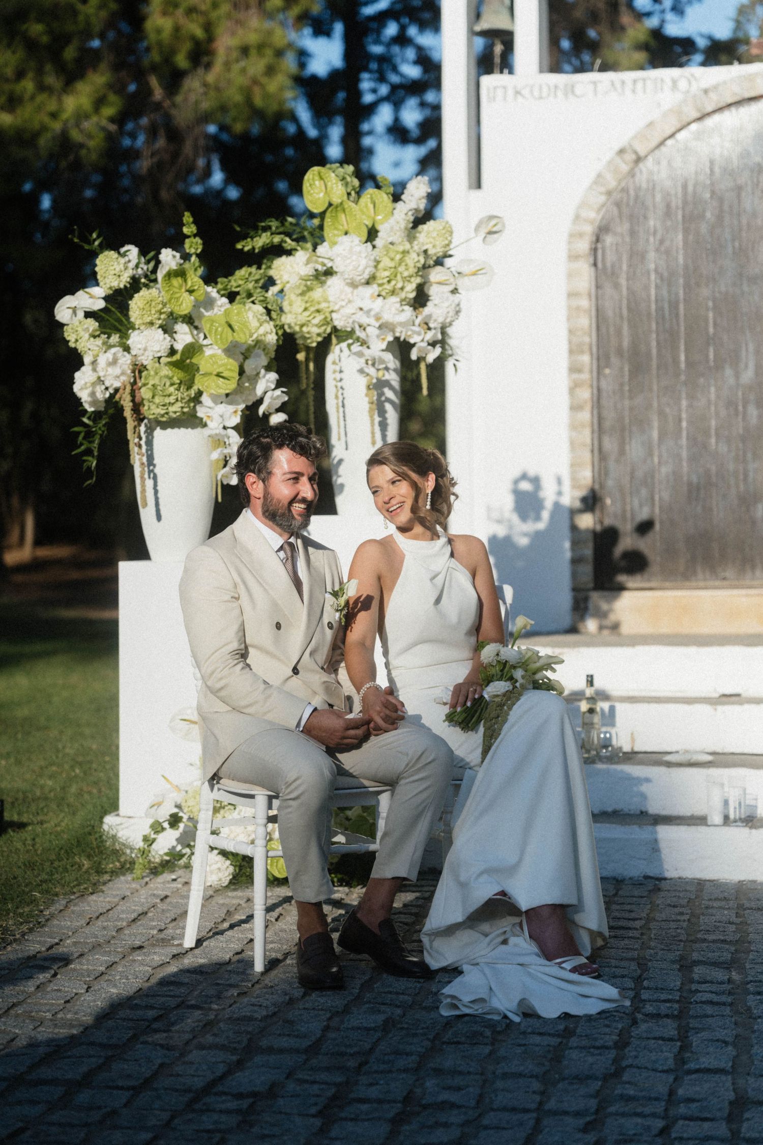 Bride and groom sitting together on white chairs at outdoor wedding ceremony with white floral arrangements.