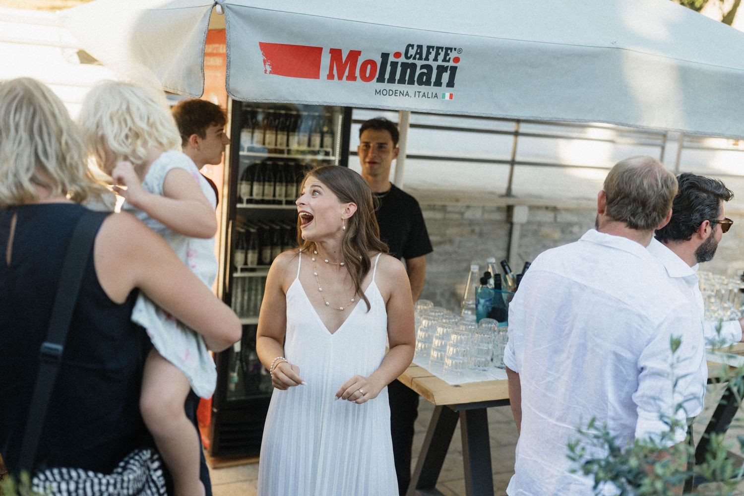 Woman laughing joyfully at outdoor Caffe Molinari coffee bar event with guests socializing in background.