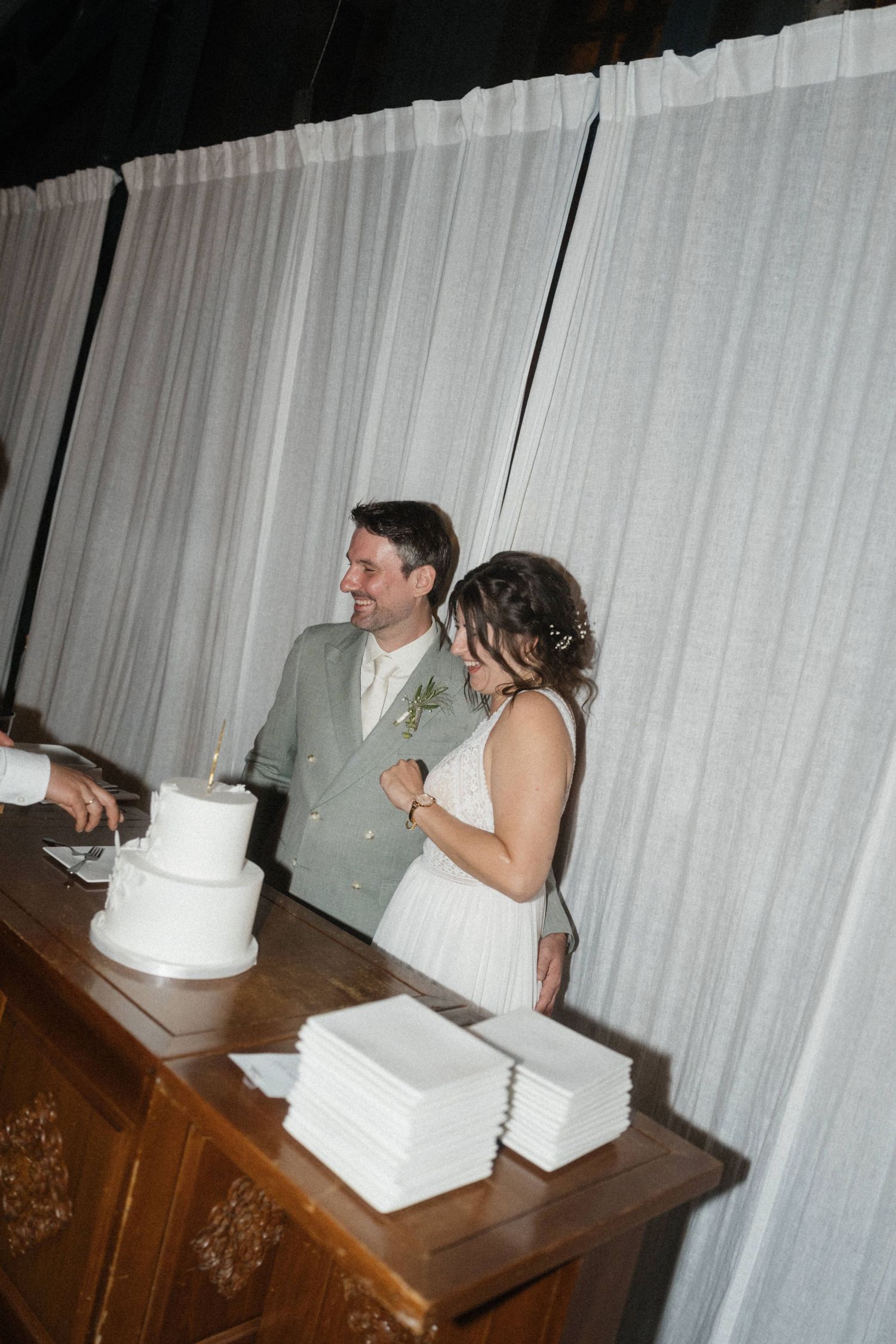 Bride and groom cutting white wedding cake at reception table with white curtain backdrop.