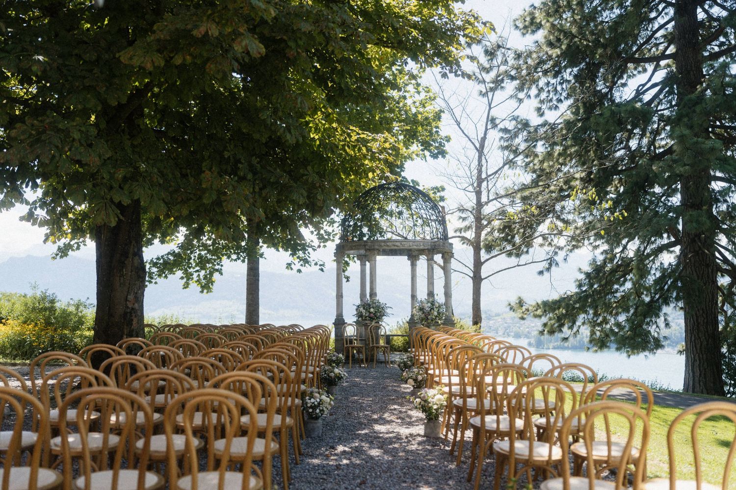Outdoor wedding ceremony setup with wooden chairs, tree-lined aisle, and elegant gazebo overlooking a serene lake.