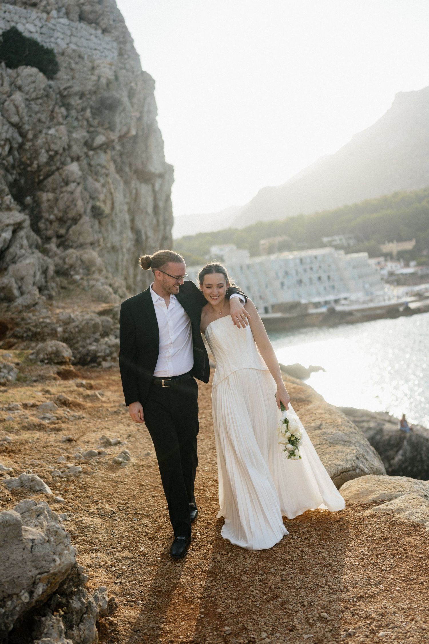 Bride in a flowing white dress and groom in a black suit walk arm in arm along a rocky coastal path with sunlit cliffs and sea behind them.